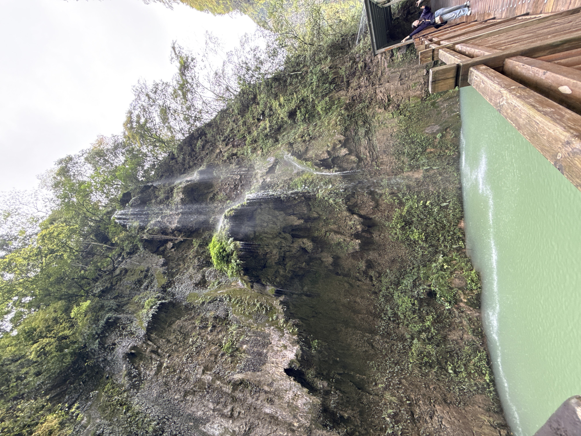 The image captures a scenic view of Baofeng Lake Waterfall in Zhangjiajie, China. A waterfall cascades down a steep, moss-covered rock face. The lake, a pale green color, lies at the base of the waterfall. A wooden viewing platform is located on the right side of the image, and two people are standing on the platform. The environment is lush with trees and greenery. The sky is overcast.