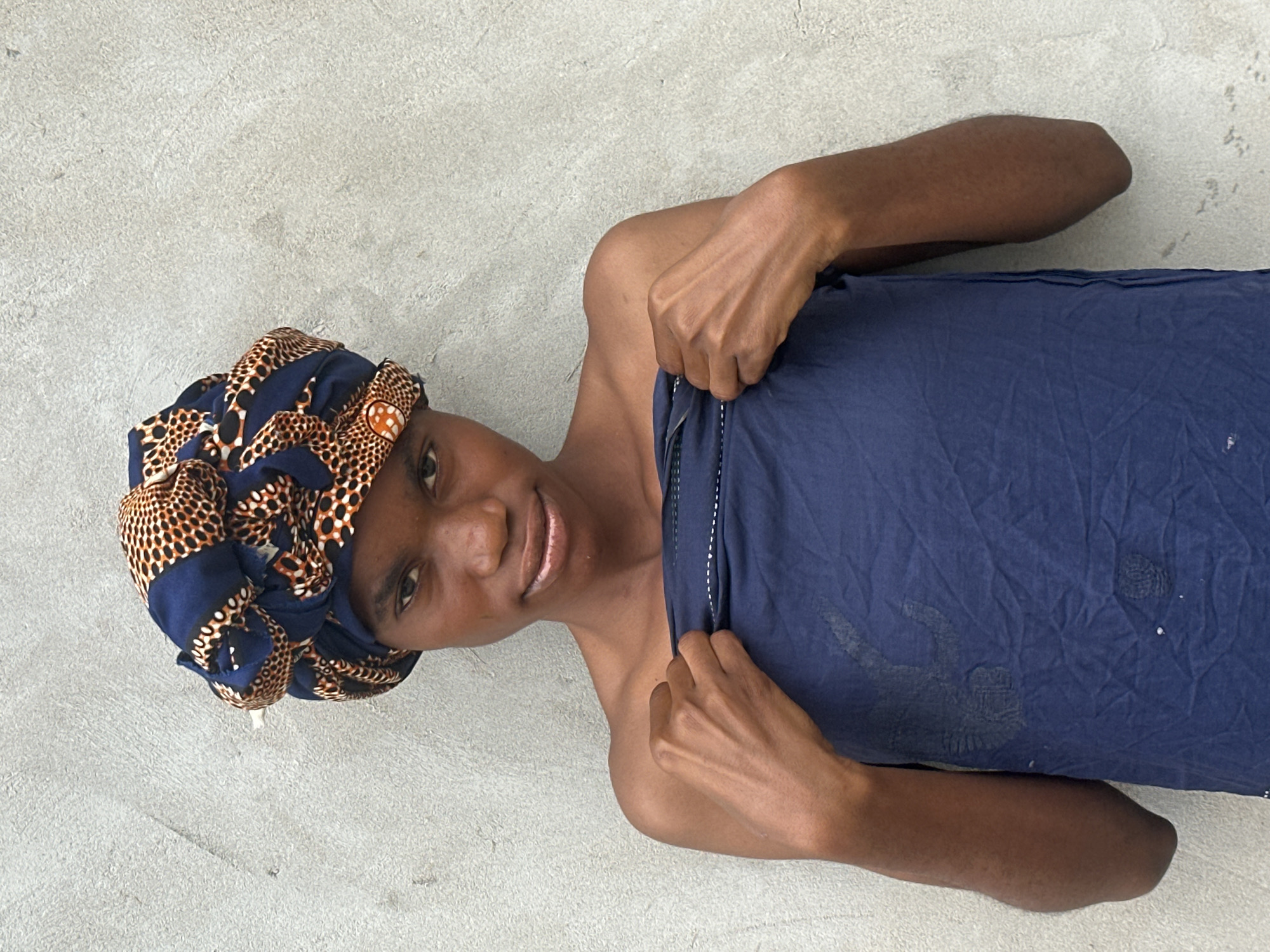 A person lies on a concrete surface, covered by a navy blue cloth. They are wearing a headwrap with a blue, black, and orange patterned fabric. The person's left arm is bent, with their hand holding the edge of the navy blue cloth. Their right arm is extended, resting on the cloth. The person's face is partially visible, and they appear to be smiling. The scene suggests a relaxed or resting posture. The concrete surface indicates an outdoor setting, possibly a yard or a courtyard, given the location context of Badagry, Nigeria. There are no visible text elements in the image.