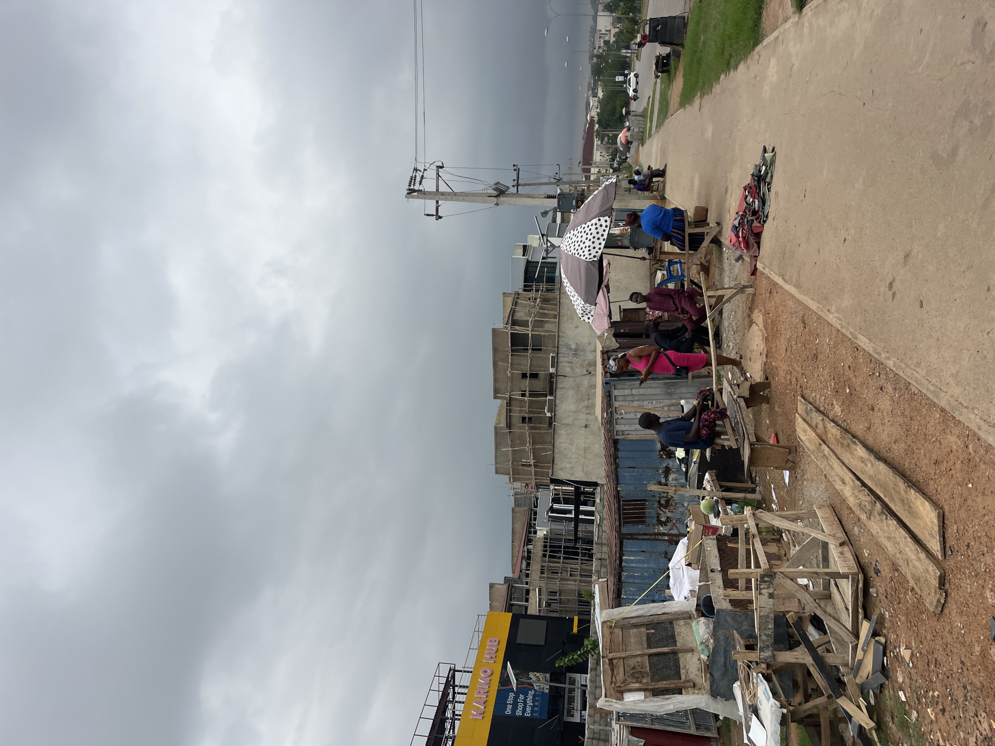 The image displays a lively roadside scene, likely in Karum, Nigeria, under a dense, overcast grey sky, hinting at cloudy weather or an approaching storm. The perspective captures a stretch of road or path lined with informal businesses and structures.
To the left, a large building features a prominent yellow sign with the text "KIARKO HUB" and "One Stop Shop For Everything" underneath, indicating a commercial center. Adjacent structures vary in completion, with some showing visible scaffolding. A tall utility pole with numerous power lines stands centrally, dominating the immediate background.
Along the right side of the image, a wide, unpaved dirt path parallels a paved road, which is partially visible. Several individuals are present, engaging in what appears to be street vending. A person in a blue shirt is seated beneath a polka-dotted umbrella, providing shade. Other people, including one in a maroon outfit and another in a pink top, are seated or standing around makeshift stalls crafted from wood, tarps, and corrugated metal. Merchandise, possibly clothing, is spread out on the ground and improvised tables. The area around these stalls is cluttered with wooden planks and various debris. The overall activity depicts routine daily commerce and roadside life in a bustling, semi-urban setting.