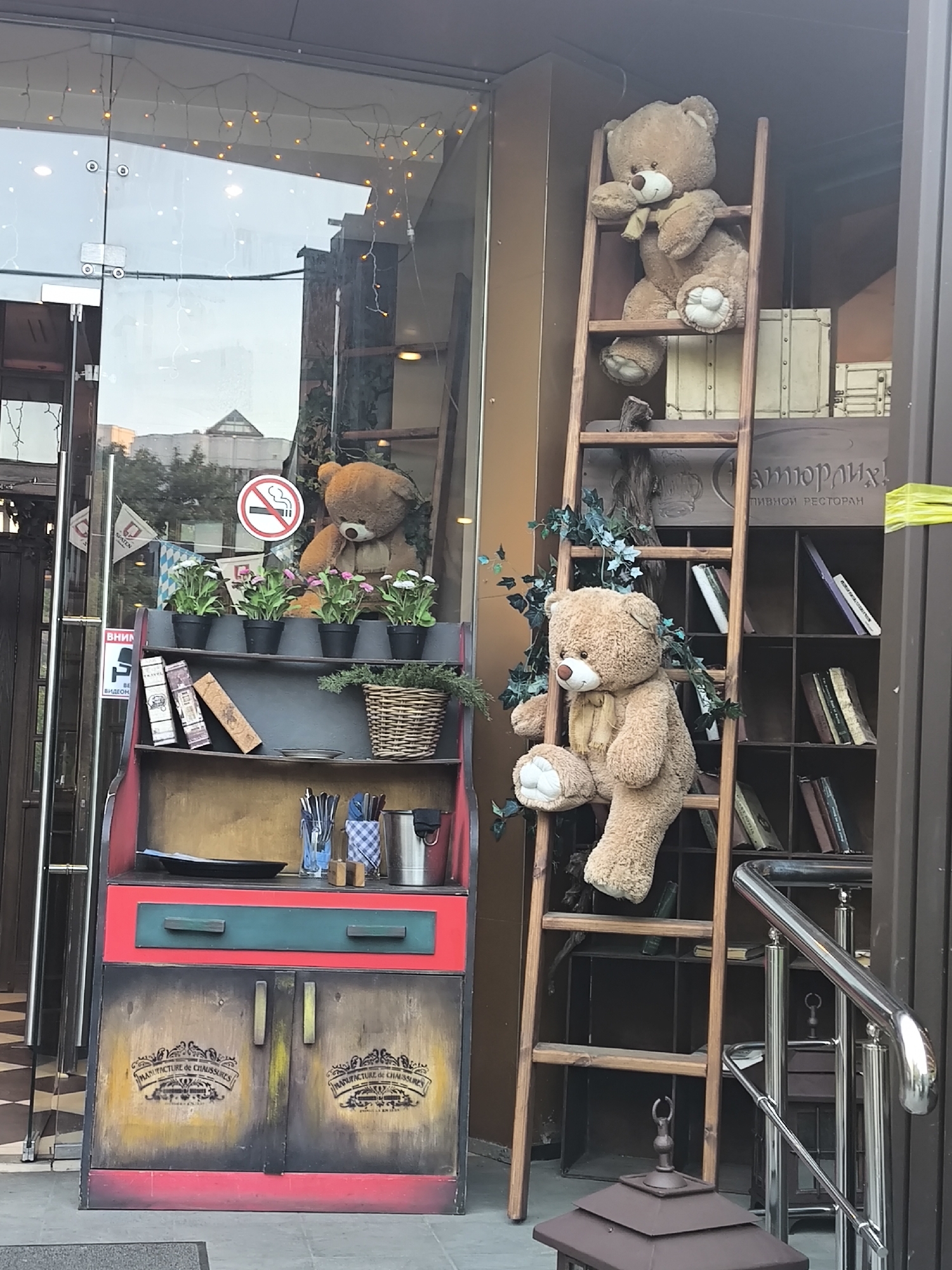 An outdoor, whimsical display decorates the entrance of what appears to be a "ПИВНОЙ РЕСТОРАН" (Beer Restaurant), likely during daytime with bright, overcast lighting. The scene features a mix of rustic furniture and plush toys, creating an inviting atmosphere.
On the left, a multi-colored cabinet-like shelving unit stands. Its top shelf holds several potted plants with small white and pink flowers, along with a few decorative books and a woven basket filled with greenery. Cutlery in various containers sits on the middle shelf. The bottom section of the cabinet has two doors, each decorated with a crest-like design and the French text "MANUFACTURE de CHAUSSURES". Behind this unit, a glass door reflects an urban environment with buildings and sky, and string lights are draped above. A red "No Smoking" sign is visible on the glass, and text reading "SPATEN" and "ВНИМАНИЕ ВИДЕОНАБЛЮДЕНИЕ" (Attention Video Surveillance) is partially visible on the doorframe. Inside, a large brown teddy bear is seen through the glass.
To the right, a wooden ladder leans against the building. Two large, light brown teddy bears with scarves are perched on the ladder; one is near the top, and the other is positioned lower down, both facing outwards. Artificial ivy with green leaves is wound around the ladder. Behind the ladder, a display of bookshelves filled with books is visible through another window or opening. The words "атюрлих!" and "ПИВНОЙ РЕСТОРАН" are legible on a sign in the background. A metal handrail runs along the far right, and a dark, ornate lantern-like object sits on the ground at the bottom right.