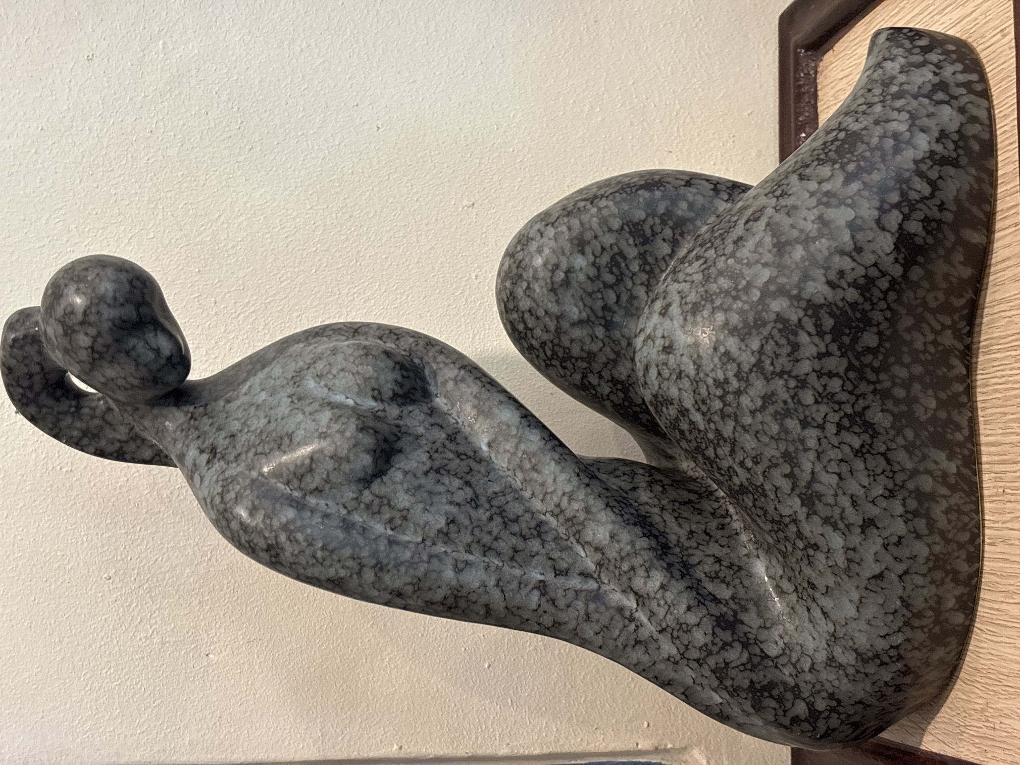 A dark gray, mottled stone sculpture of a seated female figure is displayed on a wooden base with a dark brown frame. The sculpture is positioned against a lightly textured, off-white wall. The figure is depicted in a reclining posture, with its head tilted downwards and its body curved. The surface of the sculpture exhibits a speckled texture with variations in gray and black tones. The lighting appears to be from the front and slightly from the left, casting subtle shadows. The background wall is clean and free of any visible markings or decorations, except for faint shadows. The scene does not contain any people or elements that would allow for specific location identification within Chalong, Thailand.