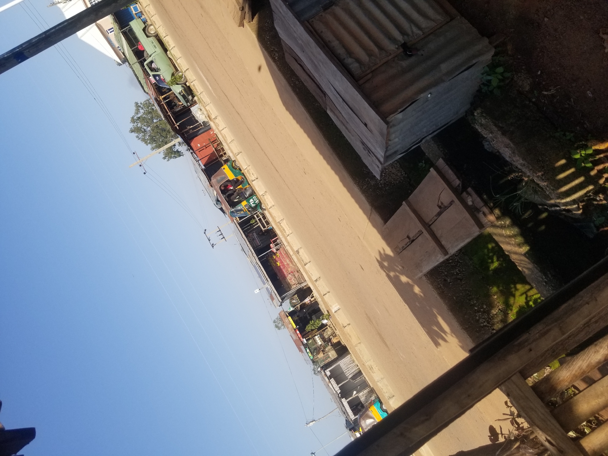 The image depicts a street scene in Jos, Nigeria, likely in a commercial or semi-residential area. The sky is clear blue, indicating daytime. In the foreground, a weathered wooden structure with railings and slats partially obstructs the view, casting striped shadows on the ground. Adjacent to this, a small, dilapidated structure made of corrugated metal stands. Beyond this, a paved road runs diagonally across the frame.
Along the roadside, several makeshift stalls or shops are visible, constructed with various materials, including corrugated metal and wood. These structures have open fronts, displaying goods or merchandise. A light green pickup truck is parked near one of the stalls, with "GOODS ONLY" written on its side.
Further down the road, a three-wheeled auto-rickshaw (Keke Napep) is stopped, with at least one person inside. The auto-rickshaw is yellow and green and has "PTA 1400" written on it. Overhead, power lines and utility poles are present, typical of urban infrastructure. Trees are visible in the background, suggesting some greenery within the urban environment. The overall impression is of a bustling, possibly informal marketplace or commercial strip on a sunny day.