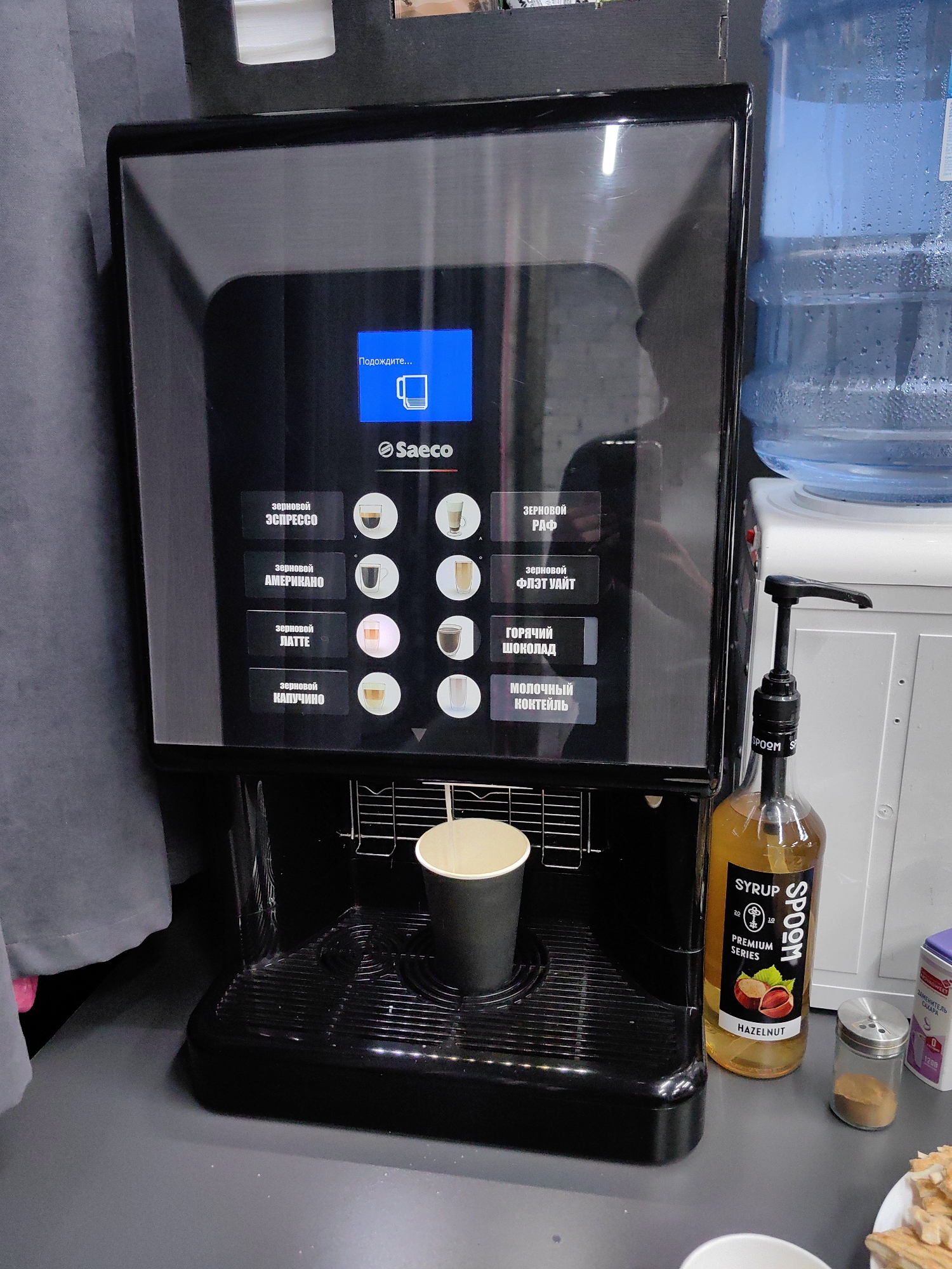 The image shows a Saeco coffee machine with a digital display showing "Подождите..." (Please wait...) and a cup icon. The machine has several buttons for different coffee drinks, including "ЗЕРНОВОЙ ЭСПРЕССО" (Grind espresso), "ЗЕРНОВОЙ АМЕРИКАНО" (Grind americano), "ЗЕРНОВОЙ ЛАТТЕ" (Grind latte), "ЗЕРНОВОЙ КАПУЧИНО" (Grind cappuccino), "ЗЕРНОВОЙ РАФ" (Grind Raf), "ЗЕРНОВОЙ ФЛЭТ УАЙТ" (Grind flat white), "ГОРЯЧИЙ ШОКОЛАД" (Hot chocolate), and "МОЛОЧНЫЙ КОКТЕЙЛЬ" (Milk cocktail).
To the right of the coffee machine is a water cooler with a large blue water bottle. Below the water cooler is a white cabinet. In front of the cabinet is a bottle of "SYRUP PREMIUM SERIES HAZELNUT" with a pump dispenser. Further to the right are a glass salt or spice shaker with a metal lid and a small container with a purple lid and a label that appears to be in Cyrillic script, possibly indicating sugar.
In the foreground, a black paper cup is placed on the coffee machine's drip tray. The overall environment appears to be an indoor setting, possibly a cafe or an office break room, with a gray curtain visible on the left. The image is taken from a medium-height angle. The scene is static, with no visible people or active interactions. The location is identified as Minsk, Belarus.