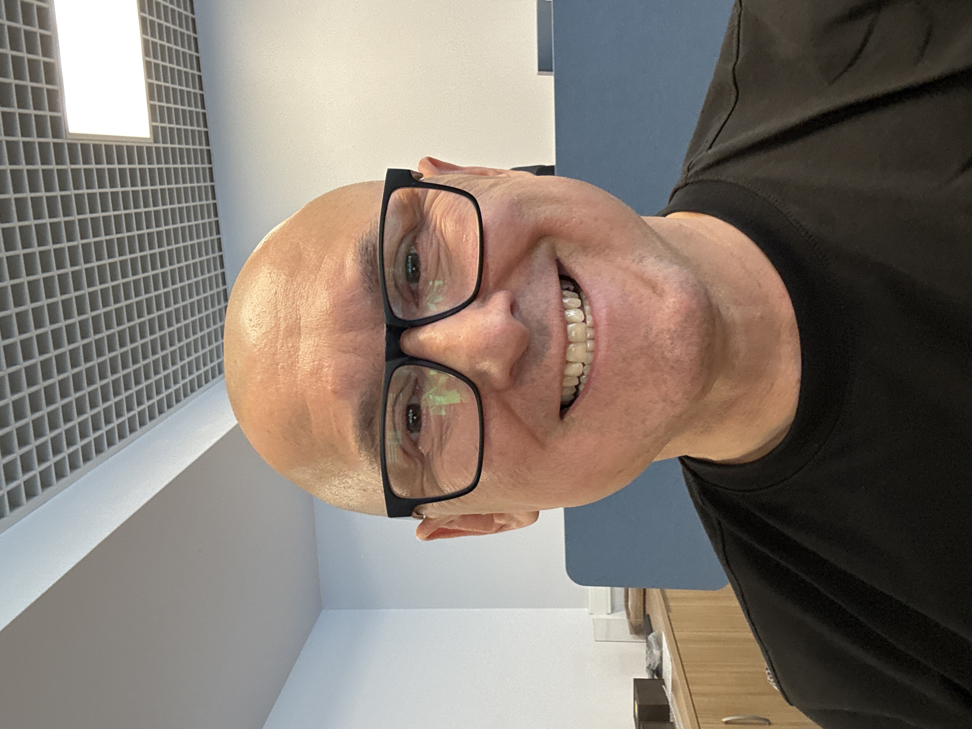 A bald, middle-aged man with glasses and a black t-shirt is looking at the camera. He is smiling and his teeth are visible. The background appears to be an office with a grid ceiling and a fluorescent light. There are cabinets with wooden drawers and a blue partition in the background. The image is a selfie, as indicated by the man holding the camera. The lighting suggests an indoor setting during the day. There are no obvious indications of the specific location within Moscow, Russia.