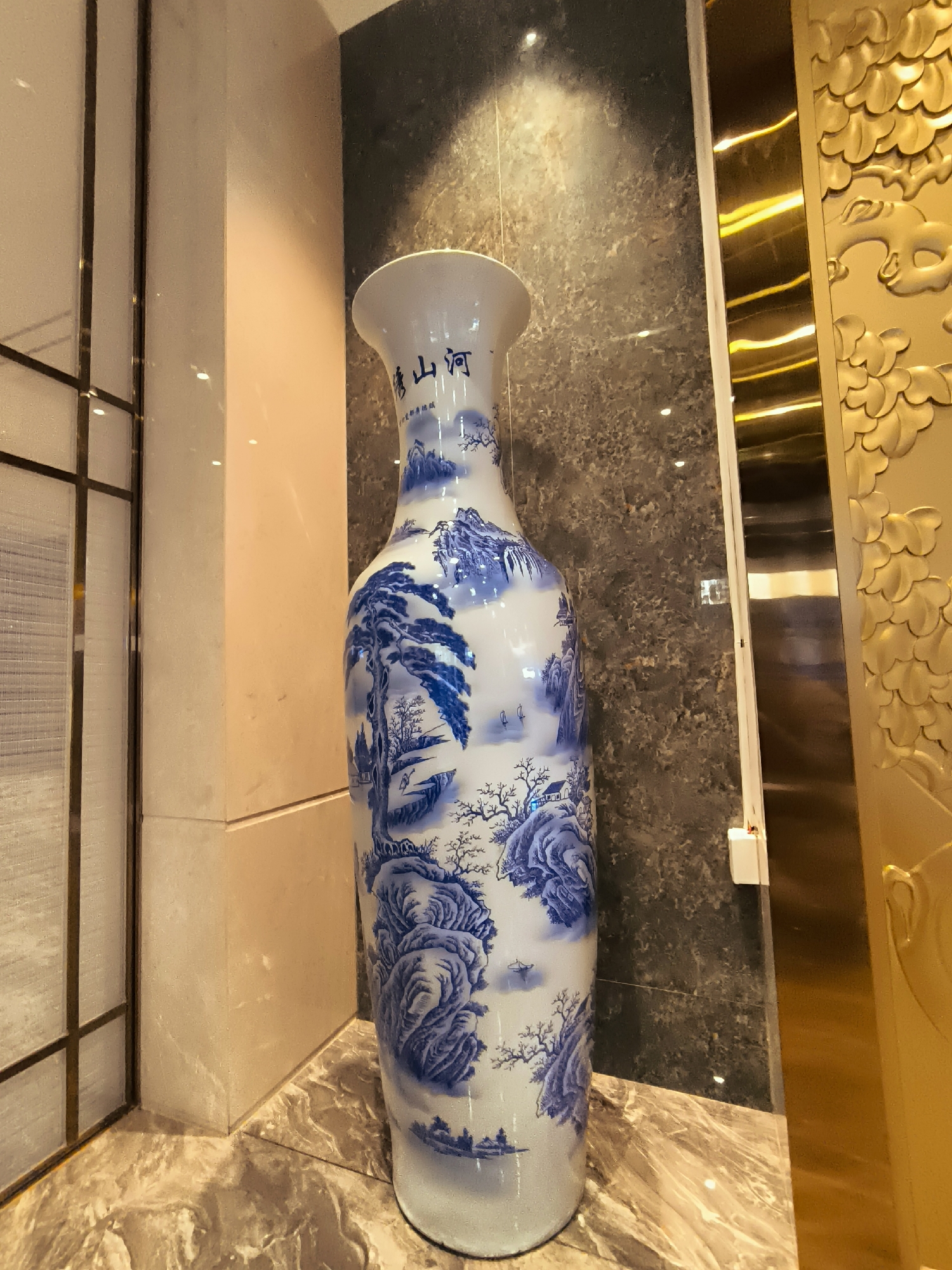 A tall, white porcelain vase decorated with blue traditional Chinese landscape paintings stands on a marble floor. The vase features intricate depictions of mountains, trees, and a pavilion, with Chinese characters inscribed near the neck. The vase is positioned in an interior setting, next to a textured gray wall and a golden decorative panel with relief carvings. To the left of the vase is a beige pillar with a vertical metallic frame. The lighting in the scene is dim, with subtle reflections visible on the surfaces. The image is captured from a medium-low angle, focusing on the vase and its immediate surroundings. The location is not discernible from the image itself.