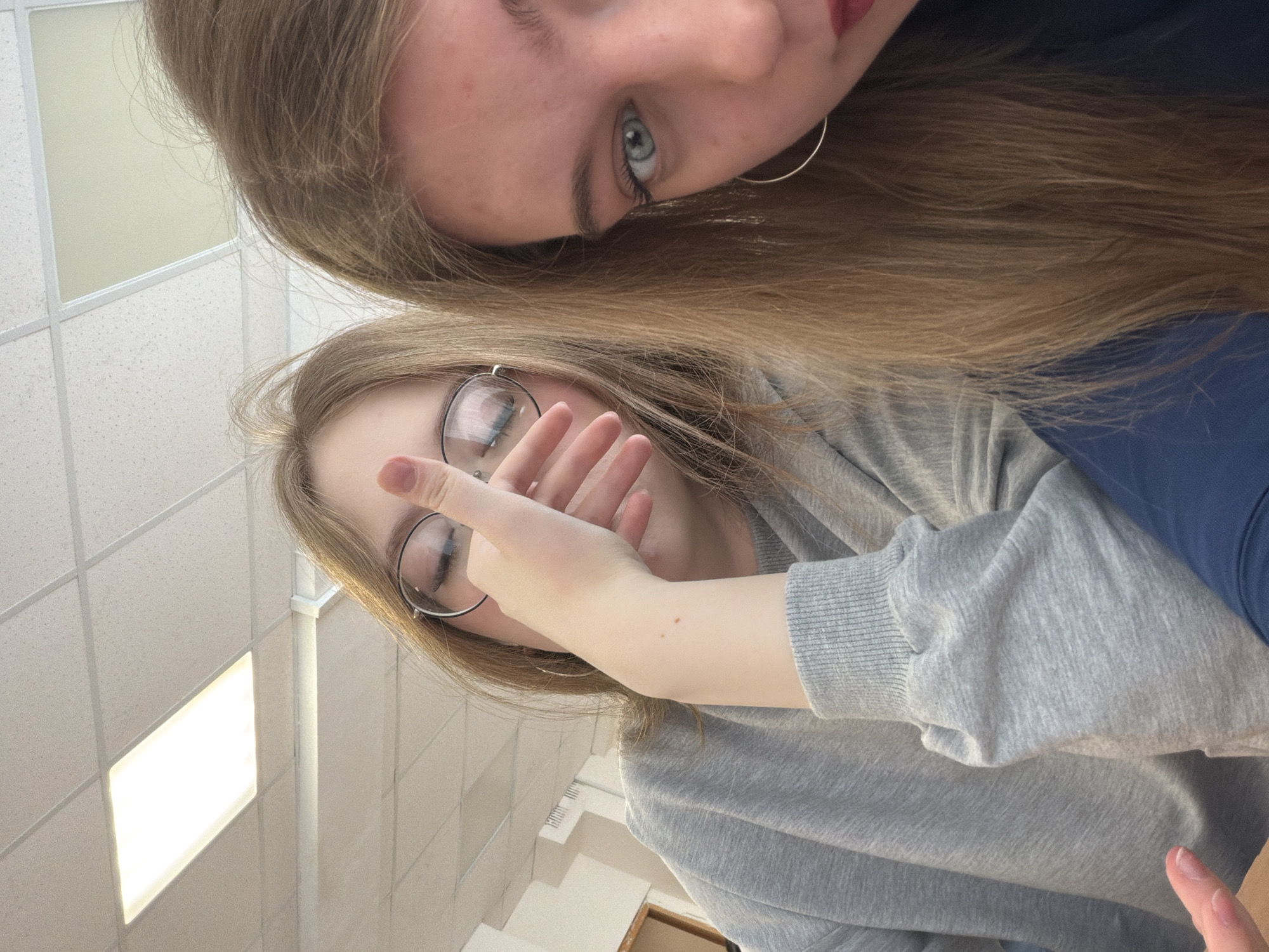 This image depicts two young female subjects situated indoors, presumably within the city of Chaikovsky, Russia. The subject on the left, with long light-brown hair, wears round-rimmed eyeglasses and a grey long-sleeved sweatshirt. Her right hand is positioned in front of her face, partially obscuring her mouth and chin. A silver hoop earring is visible on her left ear. The subject on the right, closer to the camera, shows only the right side of her face, featuring a blue/grey eye directed forward, red lipstick, and a silver hoop earring. Her long, light-brown hair is visible cascading down. She is wearing a dark blue garment. The background reveals an indoor environment with a white suspended acoustic tile ceiling equipped with multiple rectangular fluorescent light fixtures, one actively illuminated. A light-colored wall with vertical grilles and a partial wooden frame is visible behind the left subject. The scene is brightly illuminated by overhead artificial light sources.