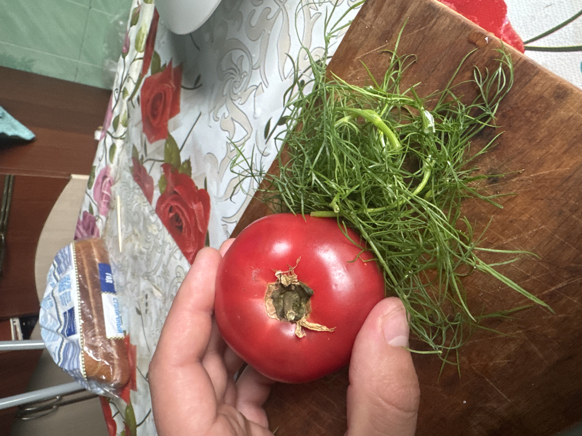 A hand is holding a ripe red tomato over a pile of fresh dill on a wooden cutting board. The cutting board rests on a tablecloth with a floral pattern featuring red roses. In the background, a loaf of bread in plastic packaging is visible on the table, along with other kitchen items and cabinetry. The scene appears to be in a domestic setting, likely a kitchen, in Красное, Russia.