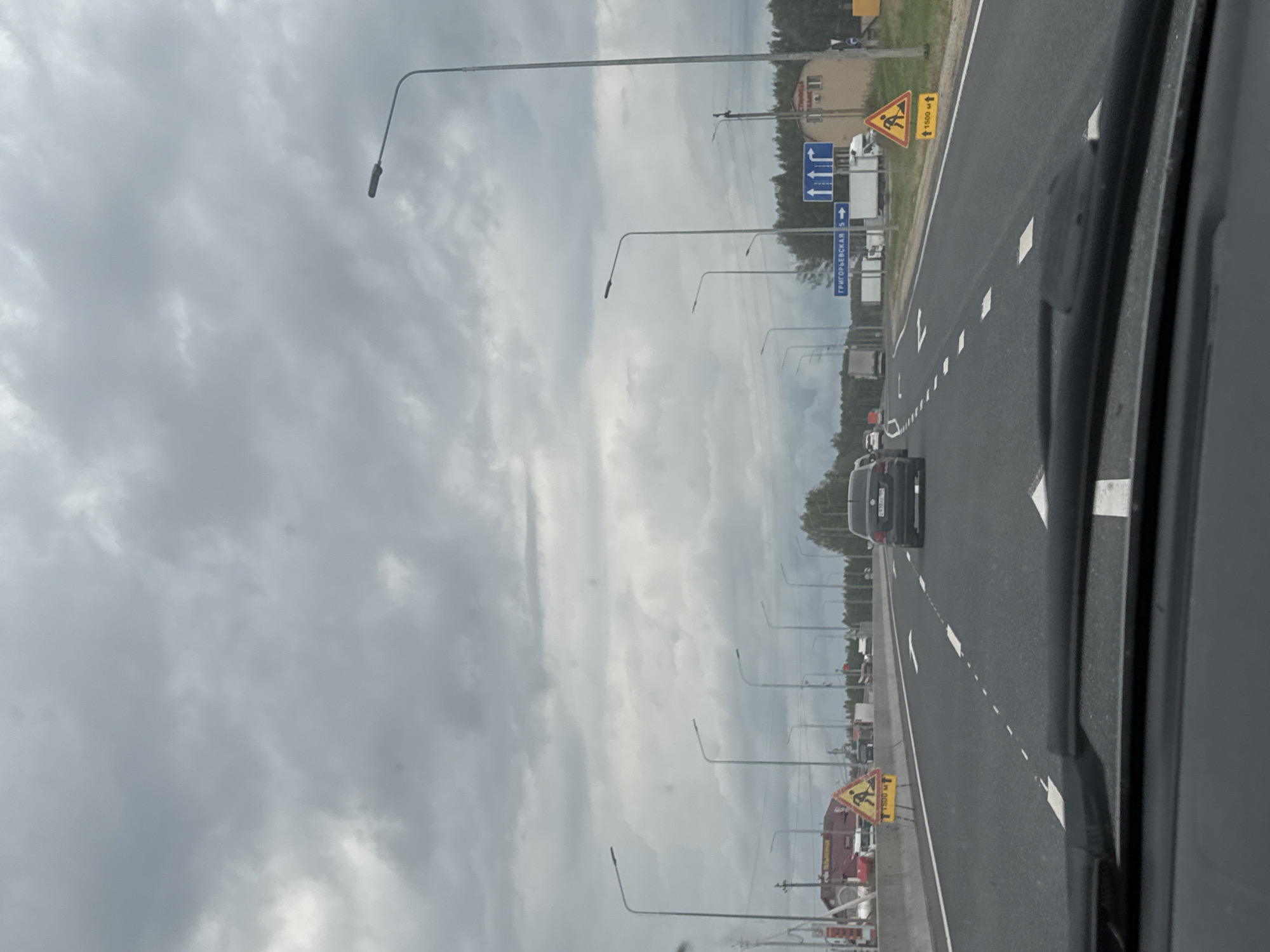 This is a point-of-view shot from inside a vehicle traveling on a multi-lane highway under a cloudy sky. The road is asphalt with white lane markings. Several other vehicles, including cars and trucks, are visible on the road. On the right side of the highway, there are streetlights, trees, a building, and road signs. One sign, a yellow triangle with a black silhouette of a person with a shovel, indicates road work ahead, with a distance marker of "1500 M". Another blue sign has Russian text and arrows indicating directions. A black Volkswagen sedan is in the right lane ahead. The location is identified as Beloborodovo, Russia.