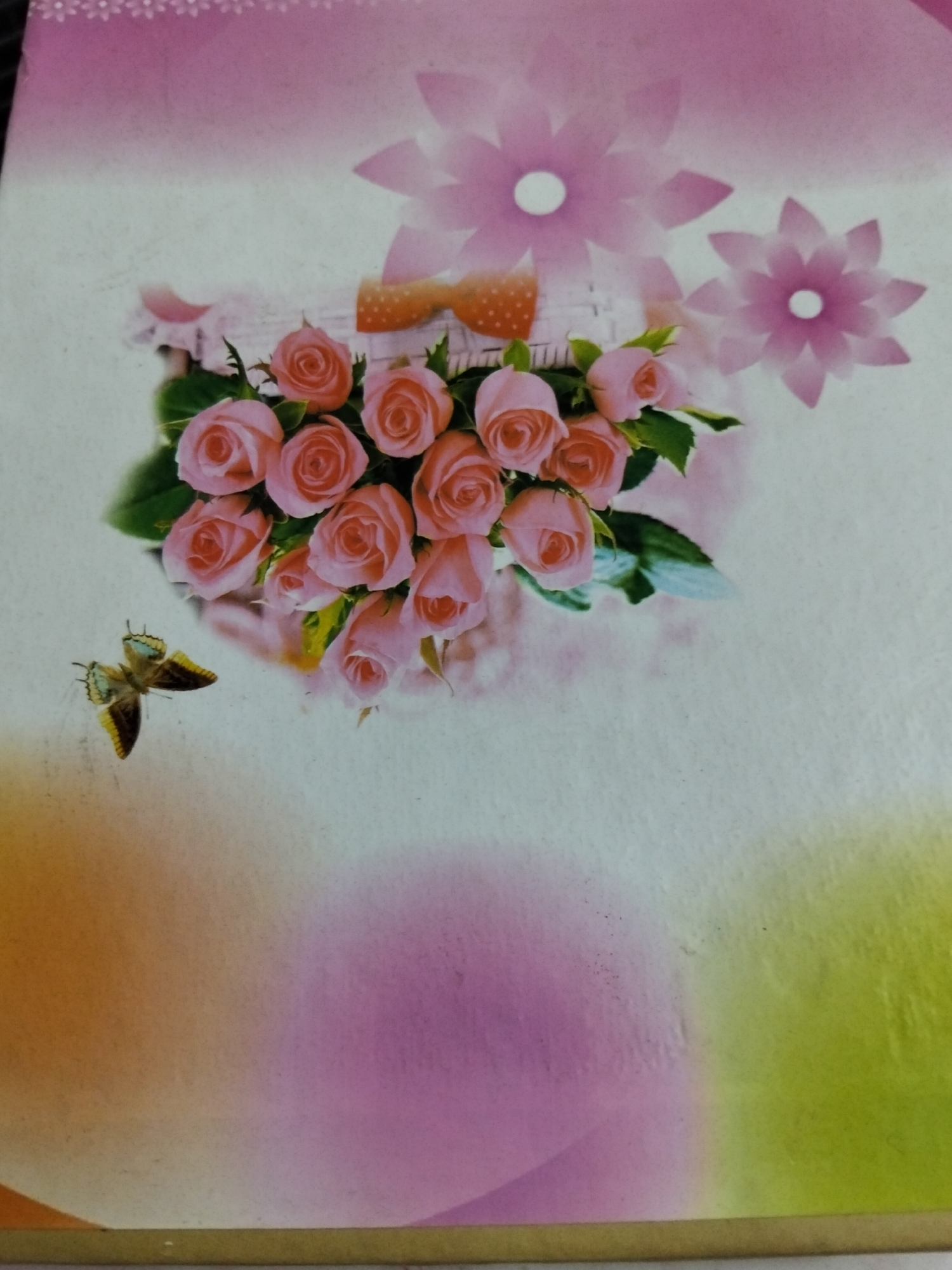 The image is a close-up of a decorative surface, possibly a notebook cover or greeting card. It features a bouquet of pink roses spilling from a woven basket, adorned with an orange bow. Two stylized pink flowers with white centers are positioned above the roses, and a small butterfly with yellow and brown wings is in the lower left quadrant. The background is a soft gradient of colors, shifting from white to pink at the top, with large blurred circles of orange, pink, and green at the bottom. There are no people or identifiable elements of Kogon City, Uzbekistan visible in the image.