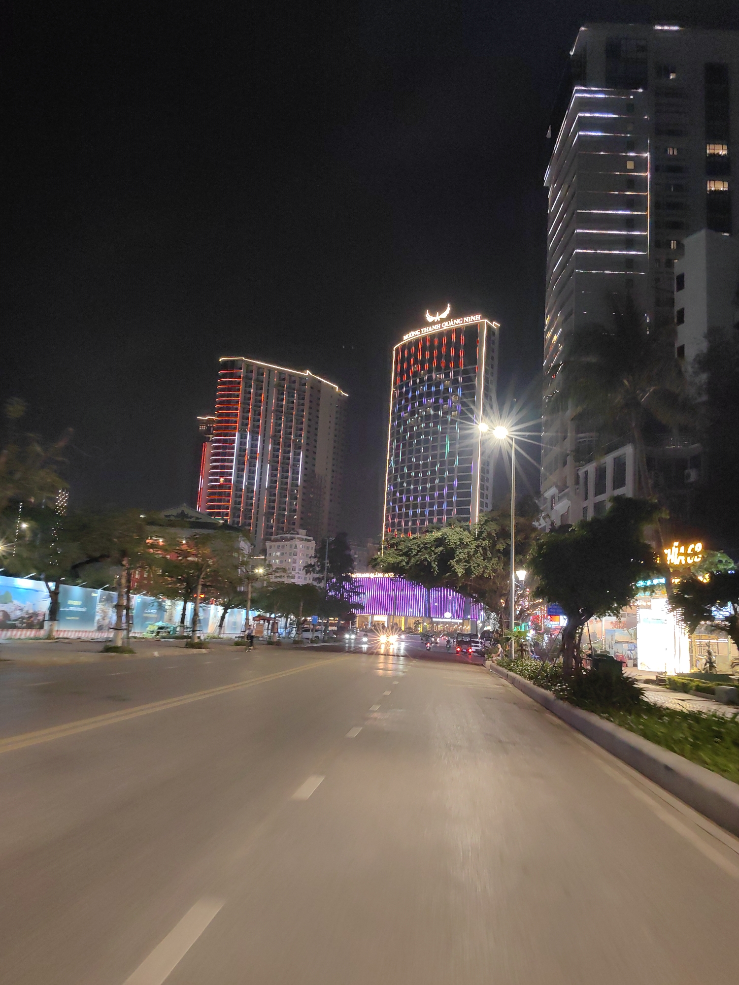 The image shows a street scene at night in Bai Chay Ward, Vietnam. The road is asphalt and appears to be wet, reflecting the city lights. There are tall buildings lining the street, illuminated with various colors. The building in the center has a logo on top. There are trees along the sidewalk on the right. Vehicles with headlights are visible on the road in the distance. The sky is dark. There are construction barriers visible along the left side of the road.