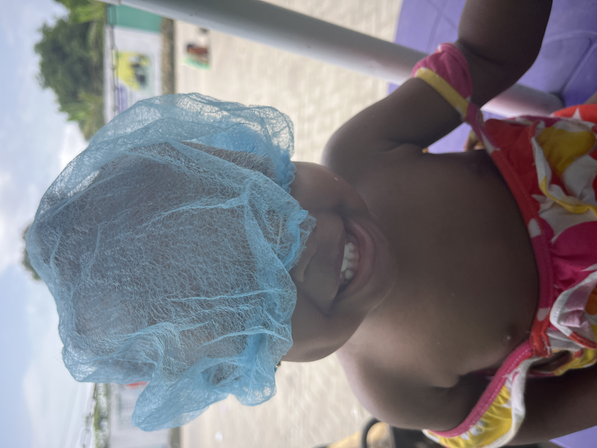 The image shows a child wearing a disposable blue hairnet and a brightly colored swimsuit. The child is smiling, revealing their teeth. The background appears to be a beach setting, possibly in Lekki, Nigeria. Vegetation is visible in the upper left corner, suggesting a coastal area. The presence of a purple object suggests furniture or seating is present. The image is slightly tilted, affecting the perspective.
