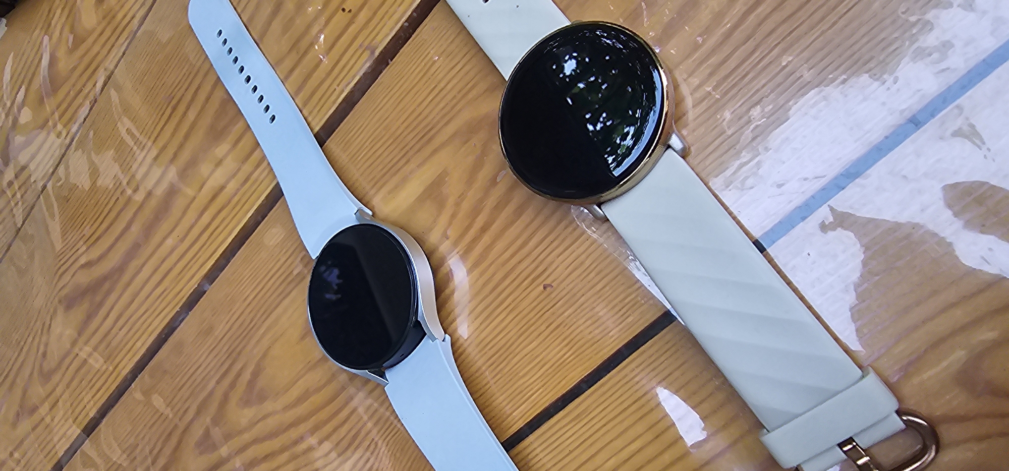 The image displays two smartwatches with light blue and pale green bands, respectively, resting on a wooden surface. The wooden surface is composed of planks with visible grain patterns. The smartwatches have round, black, reflective screens. The smartwatch with the light blue band is positioned above the smartwatch with the pale green band and slightly to its left. The pale green band of the lower smartwatch exhibits a subtle diagonal striped texture. The scene appears to be outdoors, possibly on a table or bench, given the natural lighting and the reflections on the smartwatch screens suggesting an overhead environment. There is no indication of Jizzakh, Uzbekistan, or any other specific location in the image.
