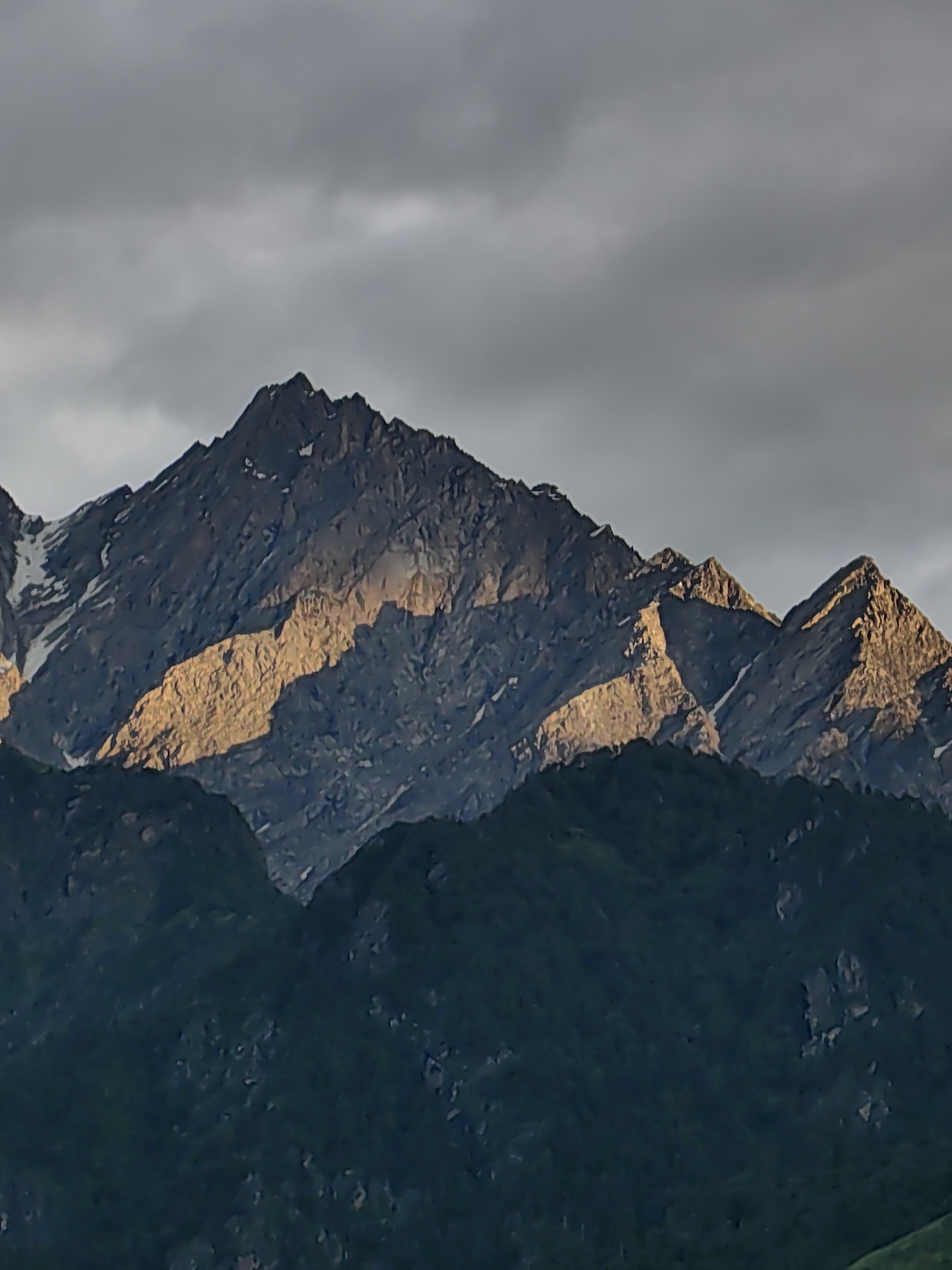 The image depicts a mountainous landscape at dusk. The sky is overcast with dark, dramatic clouds. In the foreground, there are densely vegetated, dark green hills. Dominating the background is a rugged mountain range with sharp, jagged peaks. Patches of snow are visible on the higher elevations of the mountains. A striking feature is the sunlight illuminating a section of the mountain range, casting dramatic shadows and highlighting the texture of the rock. The scene is devoid of any human presence or activity. The location is identified as Vashisht, India.
