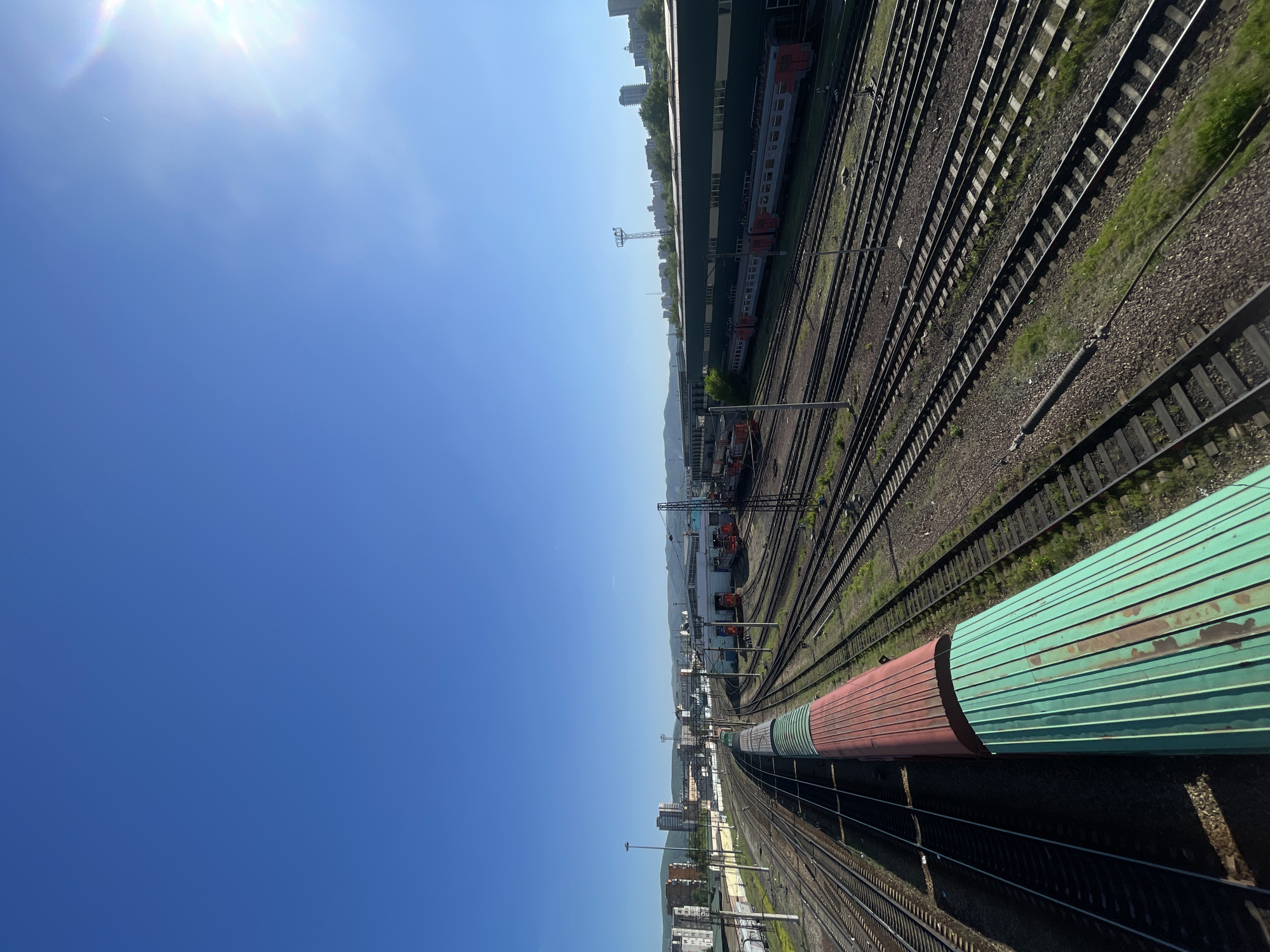 The image displays an expansive railway yard in Krasnoyarsk, Russia, viewed from an elevated perspective under a bright, clear blue sky. The sun is visible in the upper left, casting a noticeable lens flare.
The scene is dominated by numerous parallel railway tracks, some with overhead power lines and support structures, extending into the distance across the right side of the frame. In the foreground, a long train is visible on one of the tracks, featuring two carriages: one reddish-brown and the other a faded green with visible weathering and rust marks. The ground between the tracks is covered in gravel ballast with patches of green grass.
Further back, along the tracks, are several low-rise industrial buildings or train sheds. Other trains, including passenger-style carriages in grey and orange, are parked near these structures. Beyond the immediate railway infrastructure, a distant city skyline with taller buildings can be seen, backed by a range of hills or mountains under the clear sky. The bright illumination suggests it is daytime, likely mid-morning or afternoon, with no clouds present. There are no visible people or discernible text in the image.