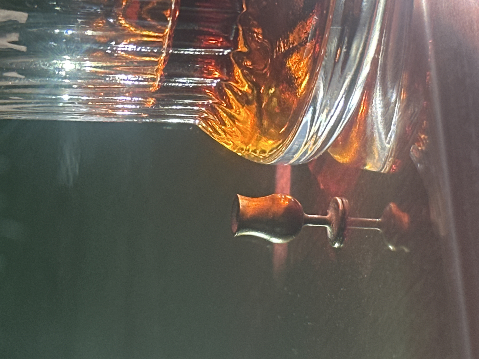 A small, dark brown, stemmed glass sits on a dark, reflective surface. To the right of the glass is a large, ribbed glass decanter filled with amber liquid. The decanter is partially illuminated, casting a warm glow and reflections onto the surface. The background is dark and indistinct, with subtle hints of green and red light. There are no people or animals visible in the image, and no indication of the specific location in Lobacheve, Ukraine.