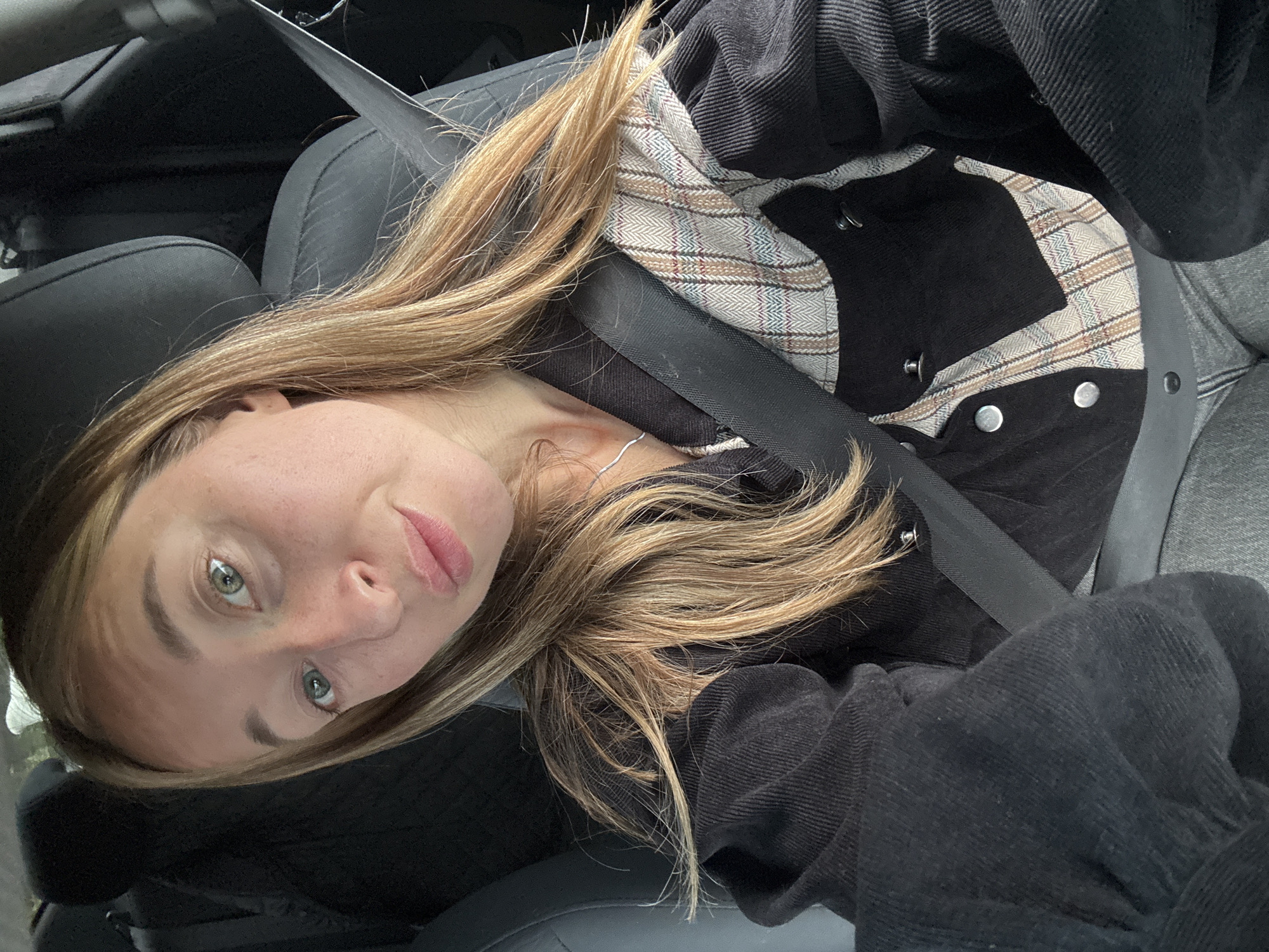 A young woman with blonde hair and green eyes is pictured from the chest up, seated in a car. She is wearing a black corduroy jacket with a plaid inner layer and a seatbelt across her chest. Her left hand is visible near her shoulder. The background shows the car's dark interior, including a seat and a headrest. The lighting suggests it is daytime. There is no discernible location within the city of Chaikovsky, Russia.
