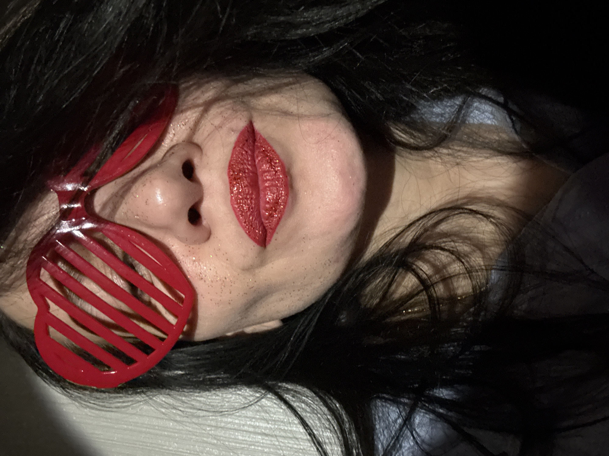 The image shows a close-up of a person's face. The person has black hair and is wearing red heart-shaped glasses. The person has red, glittery lips drawn on their cheek. The setting is unclear, with no discernible details to indicate a location in Chaikovsky, Russia.