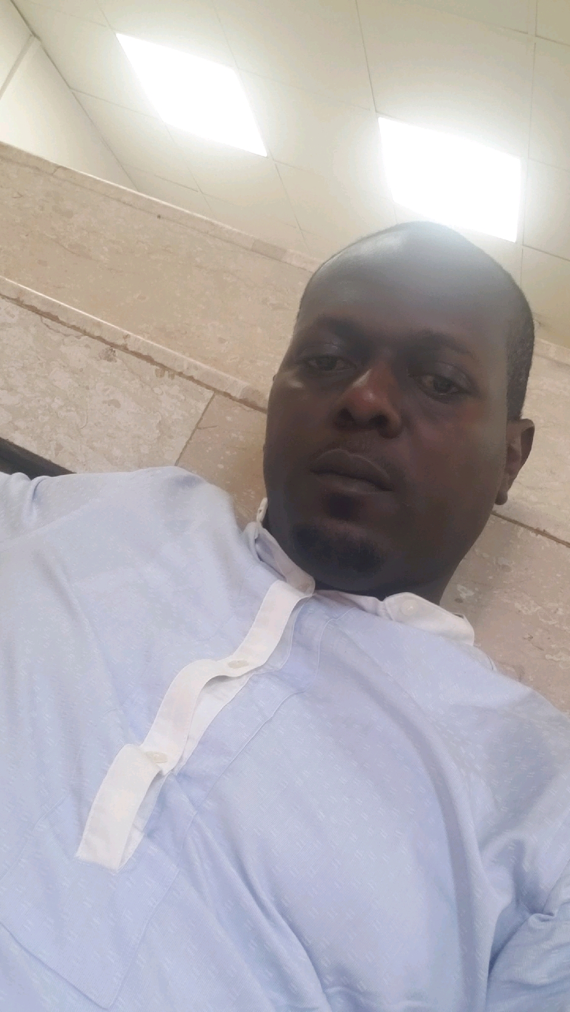 A medium shot captures a dark-skinned man with short hair and a goatee. He is wearing a light blue patterned shirt with a white collar and a white trim that buttons down the front. The man is lying down and looking at the camera. The background shows a beige tiled wall and a white ceiling with recessed lighting. The scene appears to be indoors, possibly in a residential or institutional setting in Kakuri Gwari, Nigeria.