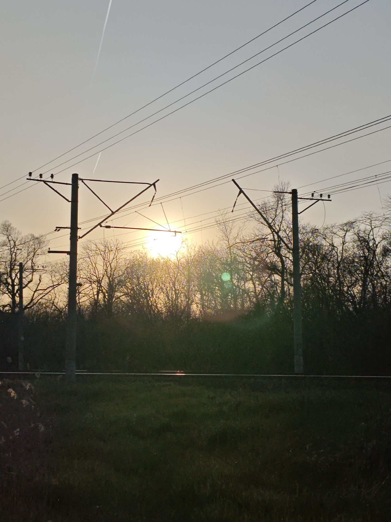 The image depicts a landscape at sunset, likely near Pyatigorsk, Russia, given the presence of overhead railway electrification structures characteristic of Russian railways. Two support masts for the electrification lines are visible in the middle ground, silhouetted against the setting sun. In the background, a dense line of trees forms a dark silhouette against the bright light of the sun. The foreground consists of a grassy field. Overhead, power lines run parallel to the ground, and a contrail is visible in the upper sky. The overall scene is a mix of natural elements and industrial infrastructure.