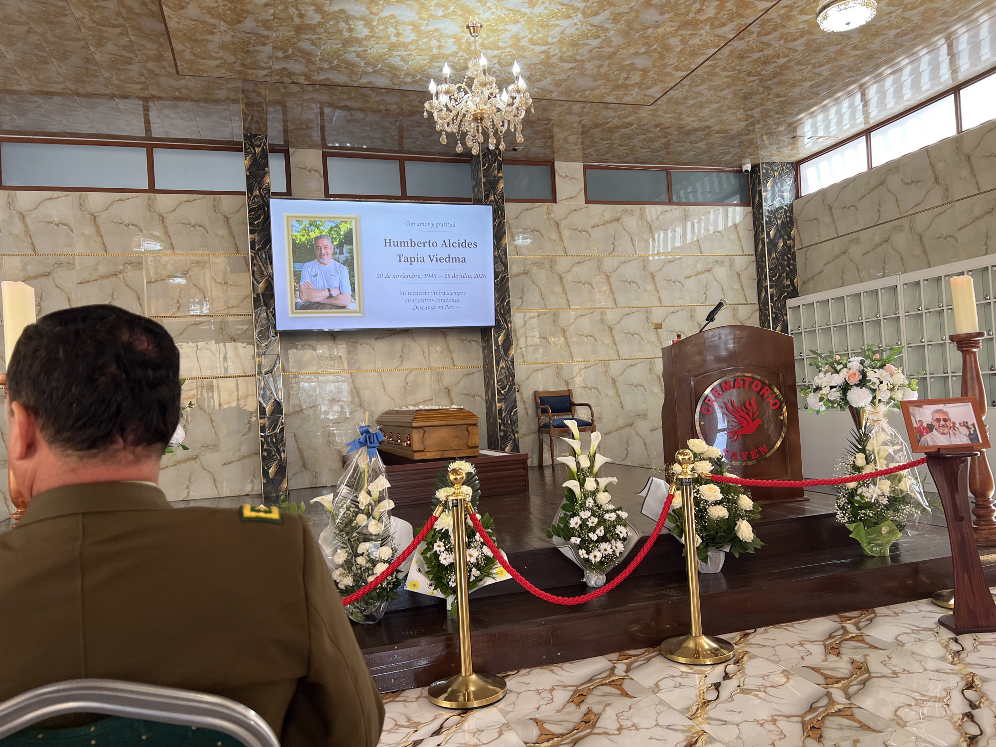 This image shows the interior of a crematorium in Arica, Chile. In the foreground, a person wearing a military-style uniform is seated, facing away from the camera. The main focus of the scene is a raised wooden platform at the rear of the room. On this platform rests a polished wooden casket, adorned with a blue ribbon. Surrounding the casket are several floral arrangements, predominantly white roses and lilies, some with clear cellophane wrapping.
To the right of the casket, a wooden lectern with a microphone stands. Above the lectern, a large screen displays a portrait of a man, identified as "Humberto Alcides Tapia Viedma," with dates indicating his birth and passing. Adjacent to the screen, a smaller framed photograph of the same man is placed on a stand, flanked by more floral arrangements. A large, ornate chandelier hangs from the intricately patterned ceiling. The walls are decorated with a marble-like finish and gold accents. Red ropes on gold stanchions cord off the platform area. The floor is tiled with a white and gold marble pattern. Natural light enters from a series of windows along the upper wall. A single large candle stands on a wooden holder to the far right.