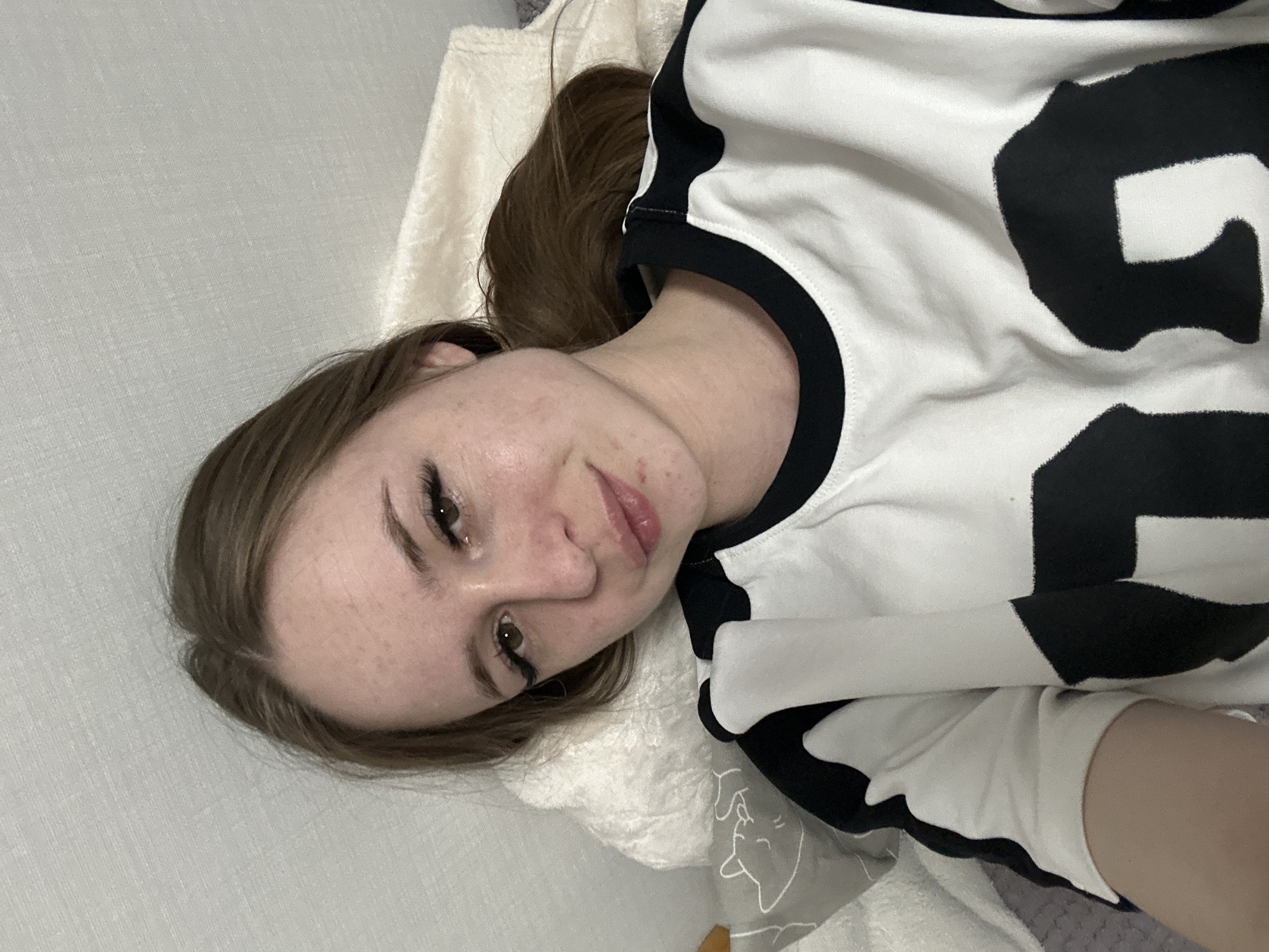 A young woman is depicted lying down, looking directly at the camera with a gentle, slight smile. Her long, light brown hair is spread around her head, resting on a light-colored, soft blanket or pillow. She is wearing a white long-sleeved top featuring bold, black, abstract patterns that resemble large, stylized block letters or shapes, with black trim around the neckline. Her brown eyes are open, and she has prominent eyelashes, possibly enhanced with mascara. Her lips are slightly parted.
The setting appears to be indoors, likely a bedroom or living room, suggested by the soft bedding and a light grey, textured wall or headboard in the background. To her right, a grey blanket with a faint outline drawing, possibly a sleeping animal's head, is visible. The lighting is soft and even, without harsh shadows. The overall activity is a casual, relaxed portrait or selfie taken while resting. No explicit text is clearly readable on her clothing, only the abstract patterns.