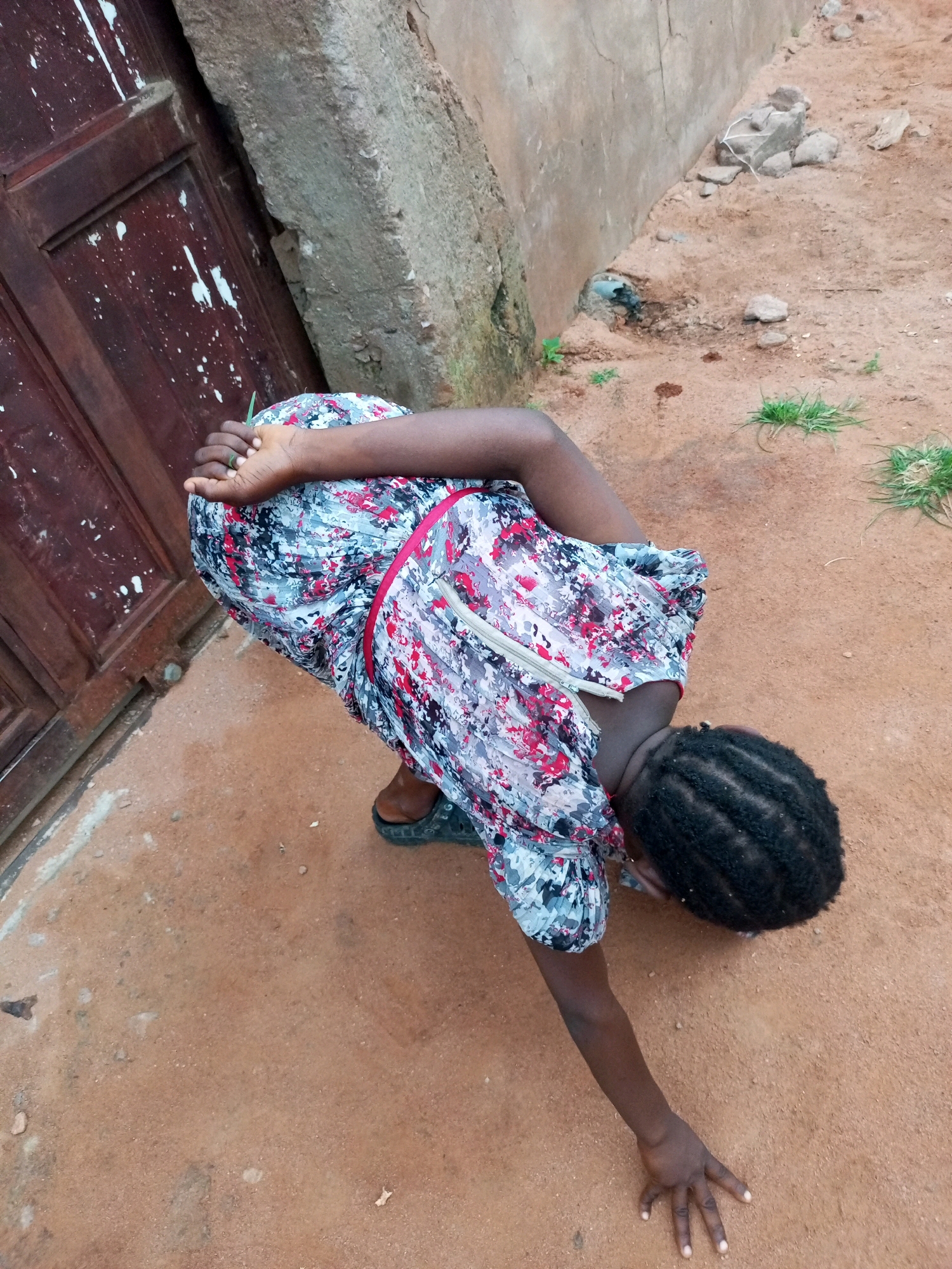 A young Black female is on her hands and knees in an outdoor dirt area, in front of a weathered brown wooden door. She is wearing a short-sleeved dress with a floral pattern of white, black, red, and blue. Her head is bowed, and her hair is in short braids. Her right arm is extended to the ground, with her hand open, fingers splayed. A green ring is visible on her left ring finger. The ground is sandy and reddish-brown, with scattered rocks and a few patches of green grass. A rough concrete wall runs along the left side of the frame, adjacent to the wooden door. The scene appears to be in a rural or semi-rural setting, possibly in Tempure, Nigeria.