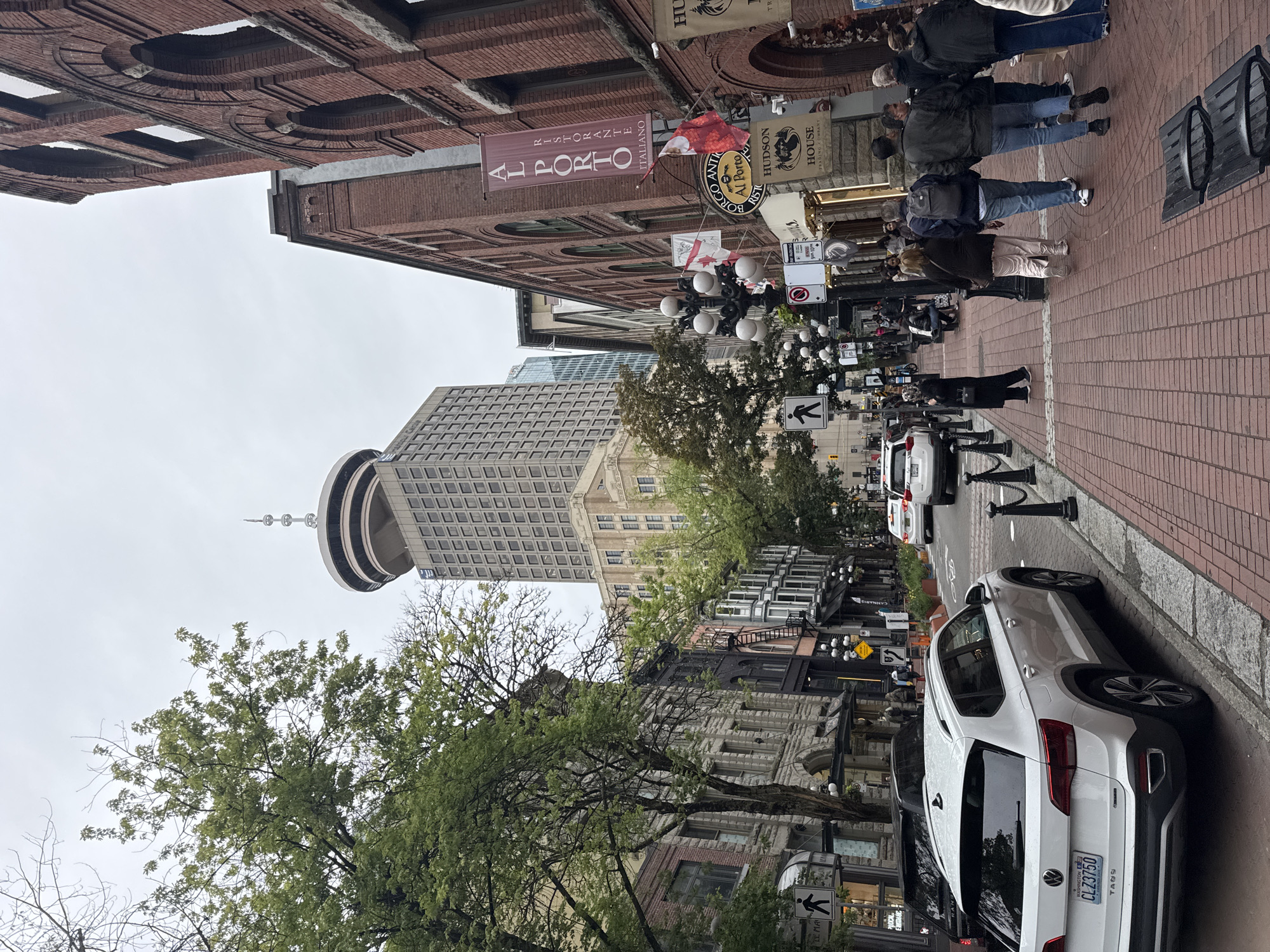 The image depicts a street scene in Gastown, Vancouver, Canada, on a cloudy day. The foreground shows a white Volkswagen Taos parked on the left side of a brick-paved street. Several people are walking on the sidewalk on the right side of the street.
The street is lined with historic brick buildings. On the right, a prominent red banner reads "AL PORTO RISTORANTE ITALIANO". Several flags are visible, including Canadian flags. Further down the street, modern buildings rise, including a distinctive tower with a circular observation deck at the top, identifiable as the Harbour Centre.
Trees with green foliage are scattered along the street, some obscuring parts of the older buildings. Pedestrians, dressed in casual clothing, are seen walking in both directions on the sidewalk. Traffic is present, with cars visible on the street. The atmosphere appears to be that of a busy urban environment with a mix of historic and modern architecture.