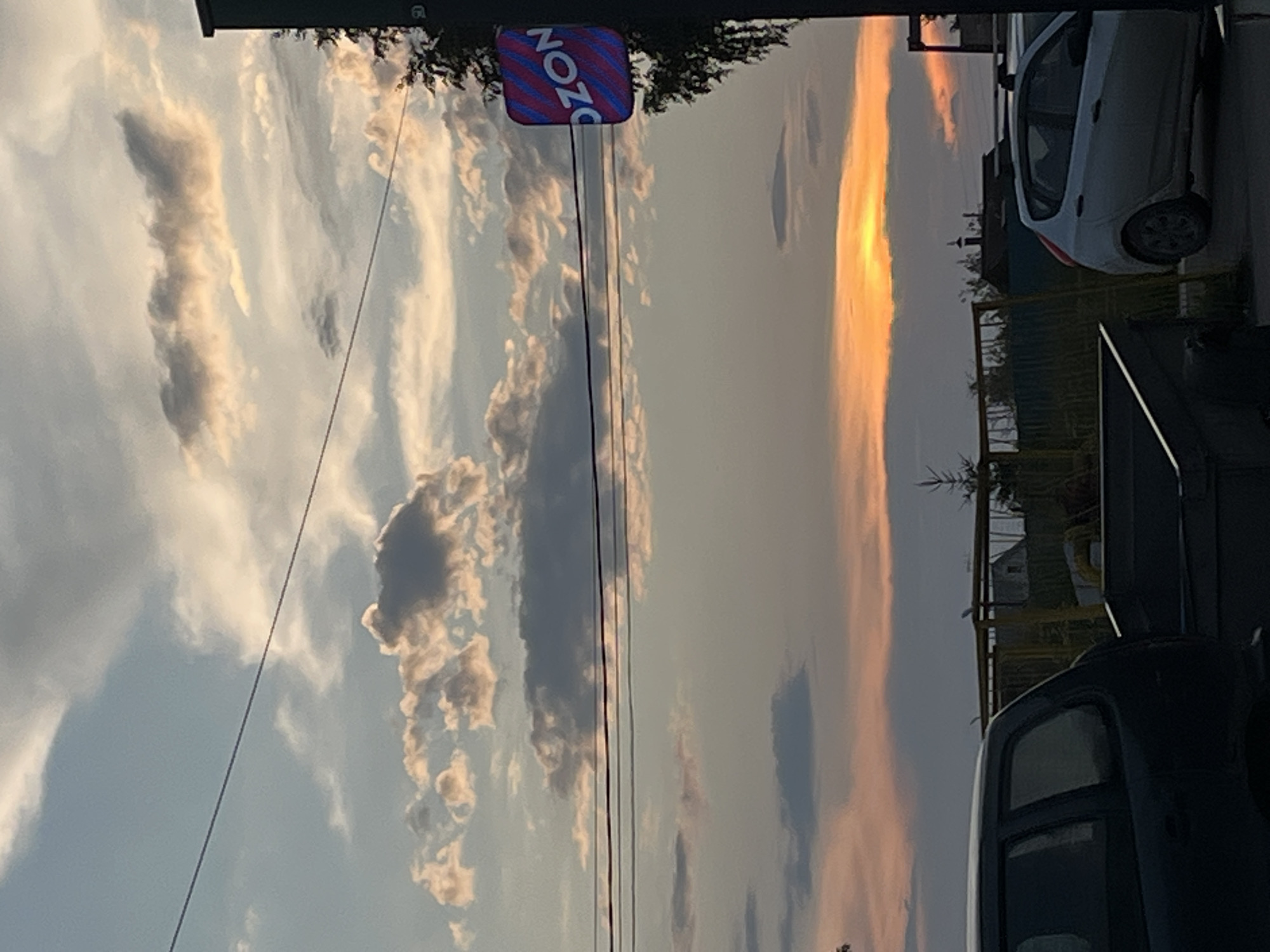 The image captures a dusky sky with scattered clouds illuminated by the setting sun, casting an orange glow on the horizon. In the foreground, a dark-colored SUV and a light-colored hatchback are parked. To the right of the vehicles, a trash bin and some yellow metal structures are visible. A sign with "OZON" written on it, illuminated with pink and blue stripes, is affixed to a pole on the right side of the image, near a tree. The scene appears to be on a street in Ludorvay, Russia, with a residential building or houses visible in the background. The overall atmosphere is calm, with the elements suggesting a typical evening scene in a semi-urban or rural setting.