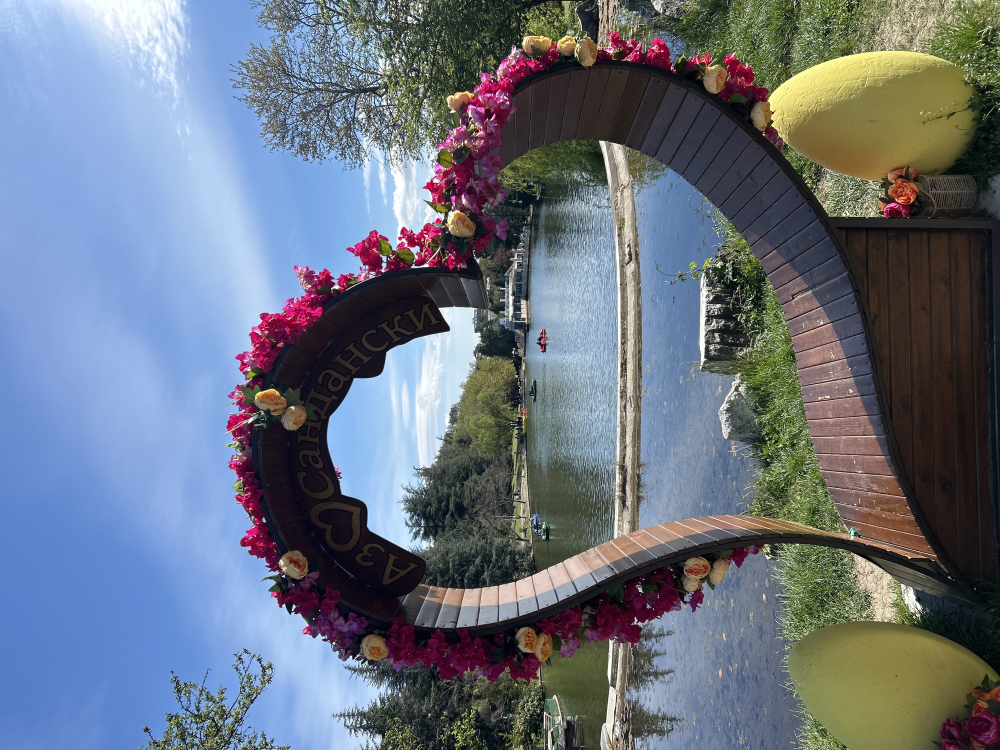 The image captures a vibrant scene in a park, presumably in Sandanski, Bulgaria, based on the Cyrillic inscription on a wooden heart-shaped structure adorned with pink and yellow flowers. The structure frames a view of a serene lake with people enjoying paddleboats or canoes. The park's lush greenery is visible in the background. A large, yellow egg-shaped object sits near the heart structure, adding to the whimsical setting. The sky is clear with scattered clouds, suggesting a sunny day.
