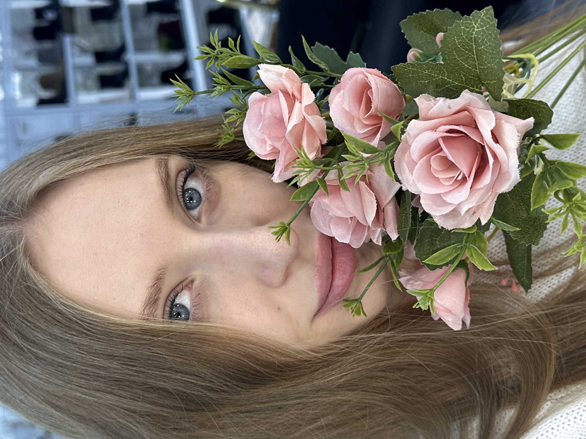 A close-up, rotated portrait captures a fair-skinned woman with light brown hair. Her eyes are blue-grey. She is positioned at an angle, with her face slightly tilted. A bouquet of light pink roses with green leaves and stems is placed near her face. The background is blurred, showing a display of products and what appears to be a knitted white textile. The location is in the city of Chaikovsky, Russia.
