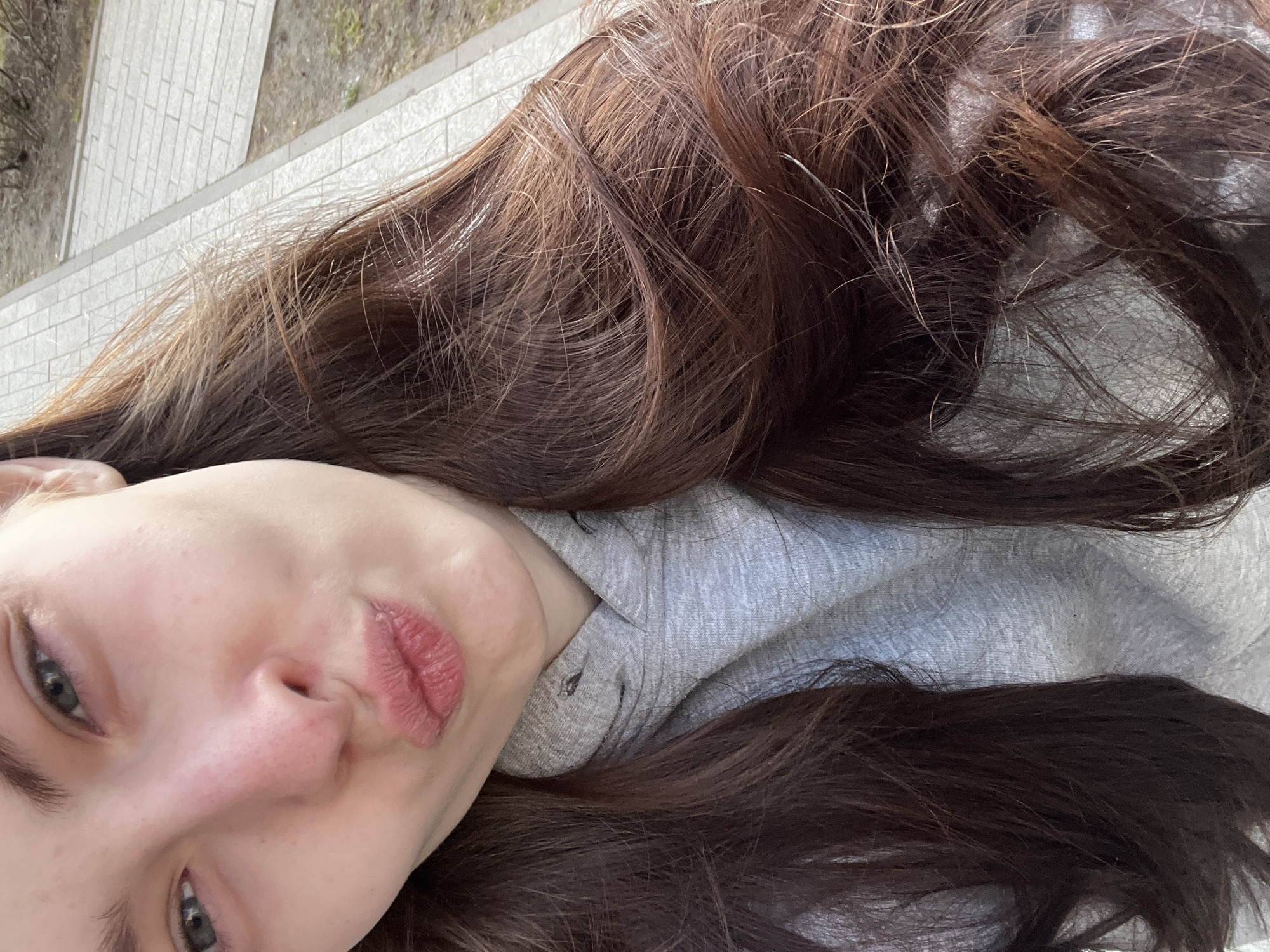 A close-up, slightly angled, high-angle shot shows a person with fair skin and long, wavy brown hair lying down, possibly outside. The person has pursed lips and visible facial features including eyes, nose, and eyebrows. The person is wearing a light gray hooded sweatshirt. In the background, there is a glimpse of a paved area that appears to be made of light-colored bricks or paving stones. The location details are not discernible to establish the image was taken in Saint Petersburg, Russia.