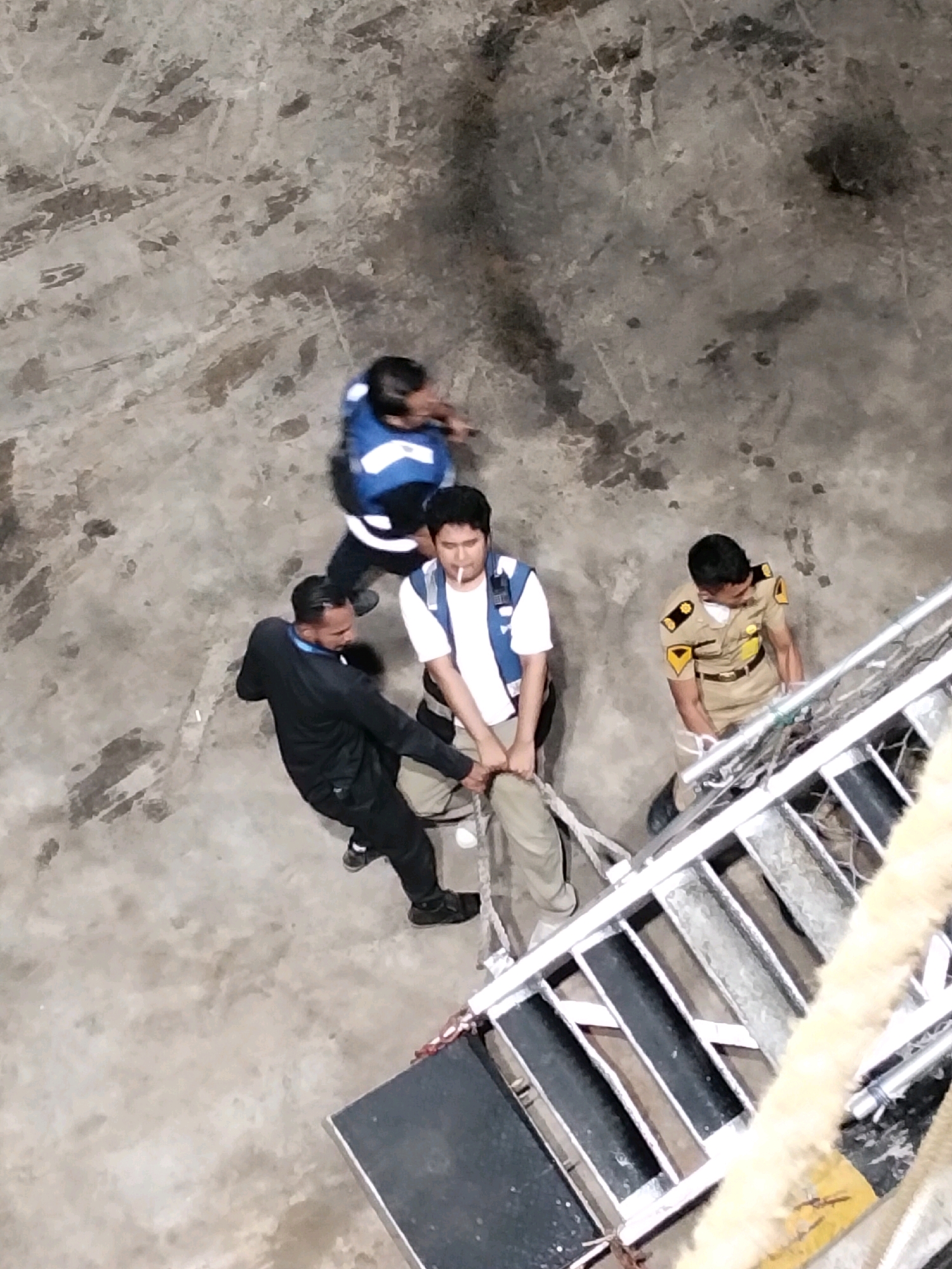A group of four individuals are on a concrete surface, possibly a dock or industrial area, in Batam, Indonesia. One person, wearing a blue vest with white accents, is walking away from the camera. Another person, wearing a white t-shirt and a blue vest, is holding a rope and appears to be interacting with another individual in a black tracksuit. The person in the tracksuit is also holding the rope, which is attached to a metal structure that looks like a ladder or ramp. A fourth individual, dressed in a tan uniform with yellow insignia on the shoulders, stands near the metal structure, looking down at it. The ground is textured concrete with some dirt or debris.