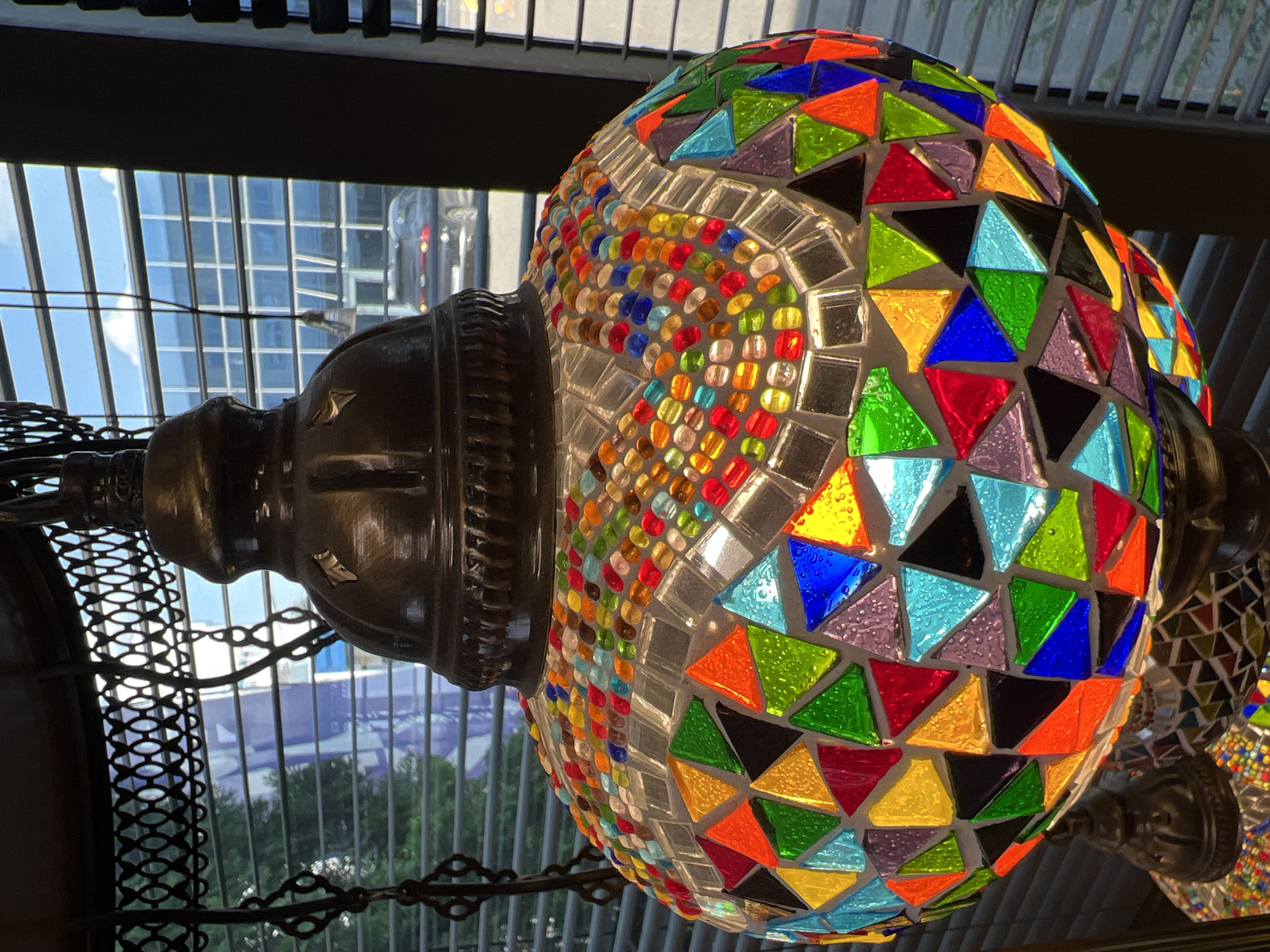 A mosaic lamp with a rounded base and a bell-shaped top is prominently featured in the foreground. The lamp is decorated with numerous small, colorful glass pieces in various geometric shapes, predominantly triangles and squares. These glass pieces are set against a white background, creating a vibrant mosaic pattern. The lamp is suspended by a dark metal chain and frame.
In the background, through a window with horizontal blinds, a modern building with a glass facade is visible. The sky above is bright blue with scattered white clouds. A car is also visible parked in front of the building. The overall scene suggests an indoor setting, likely a shop or a home, with a view of an urban environment. The image does not contain any information to suggest a specific location in Krasnodar, Russia.