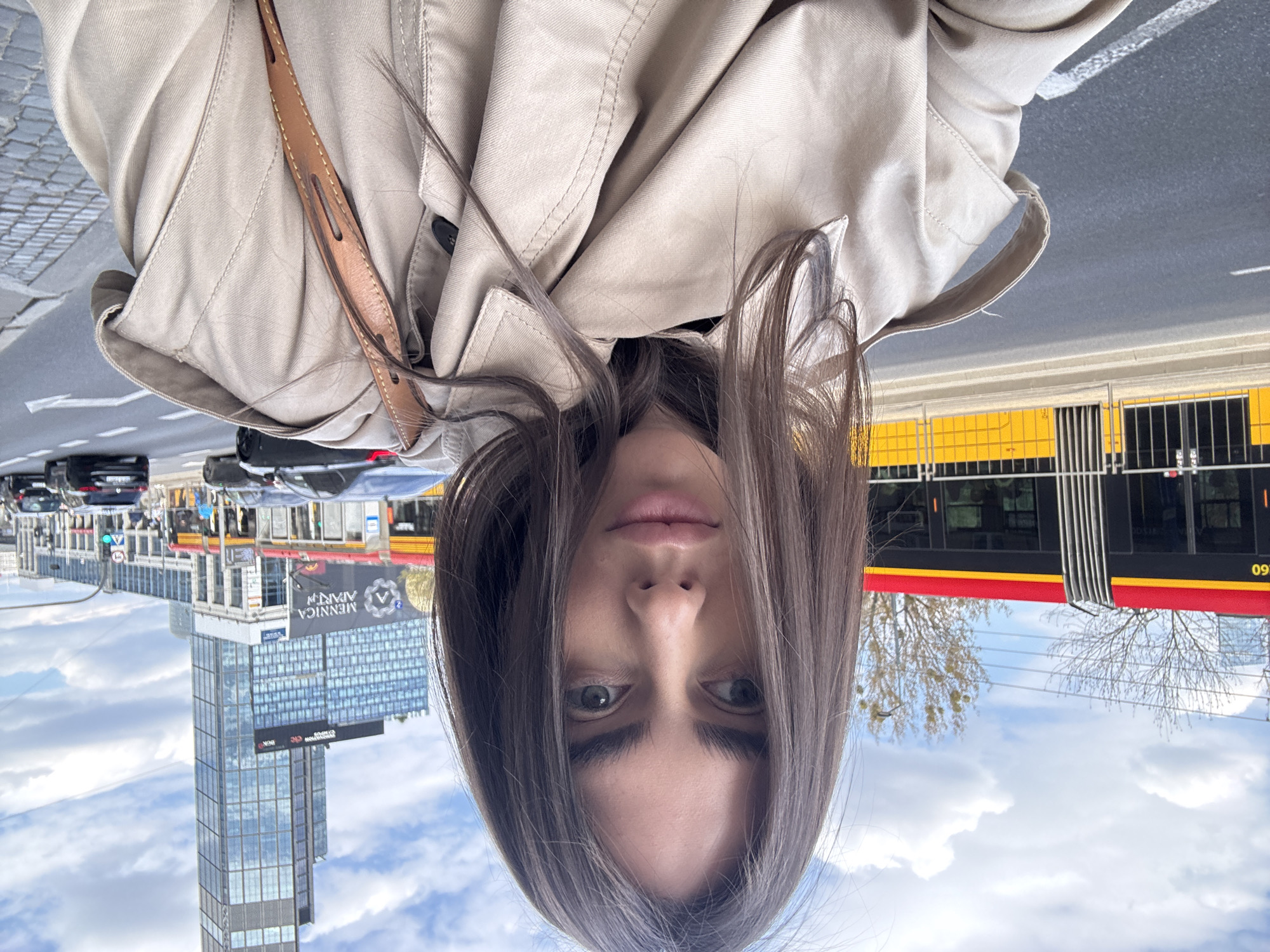 A selfie shows a woman outdoors in Warsaw, Poland. She wears a beige trench coat with a brown strap. Behind her is a city street with a yellow and red tram, cars, a skyscraper with "Mennica Apartments" signage, and a sky with clouds.
