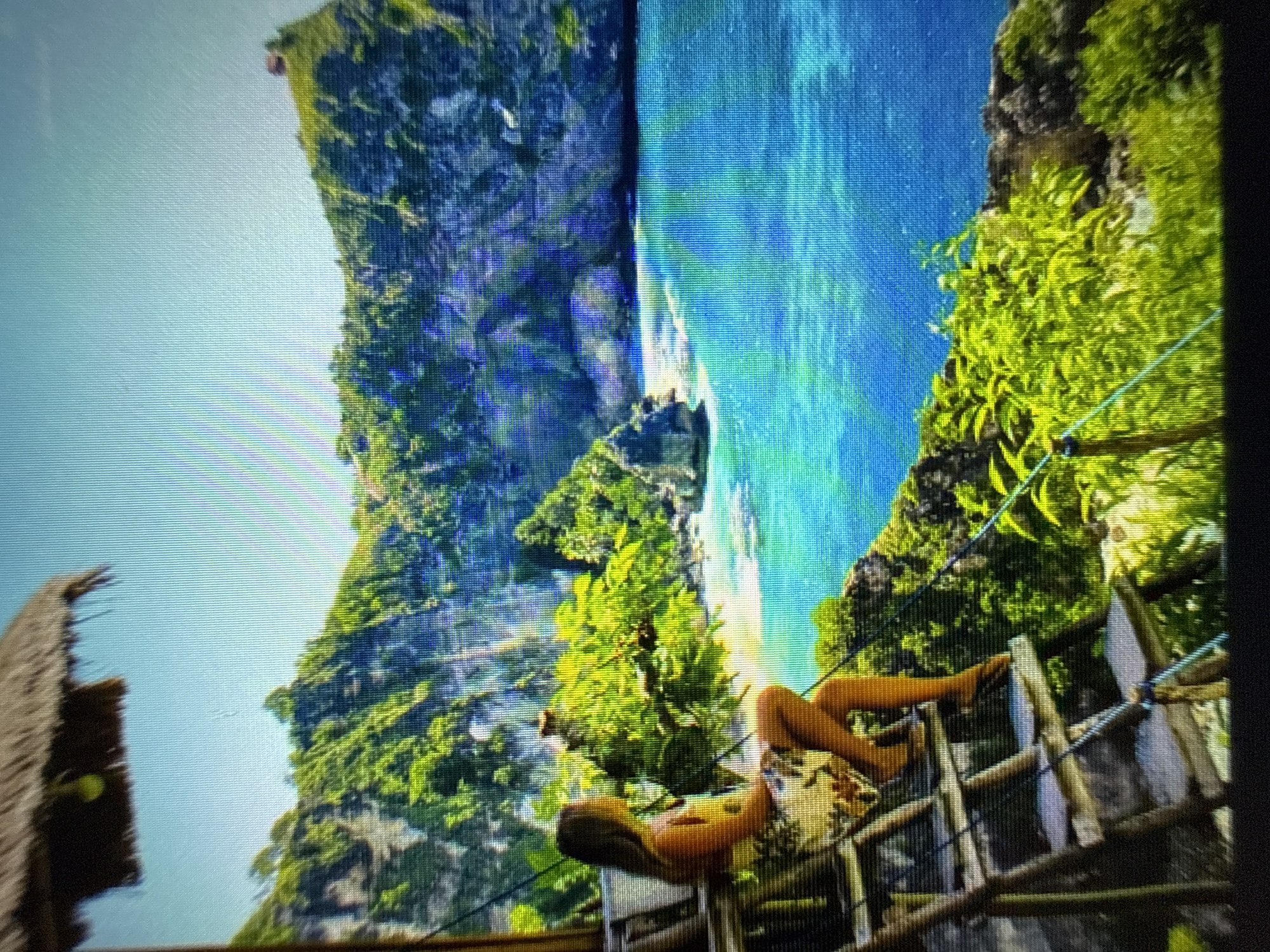 A woman sits on a wooden staircase, facing a tropical ocean vista. She is wearing a floral-patterned dress and holding a book. The staircase is constructed of rough-hewn wood and secured with ropes. Lush green foliage surrounds the staircase and overlooks the vibrant blue ocean, which meets a rocky coastline and a cliff face covered in trees. The sky is clear and bright blue. A thatched-roof structure is partially visible in the upper left corner.