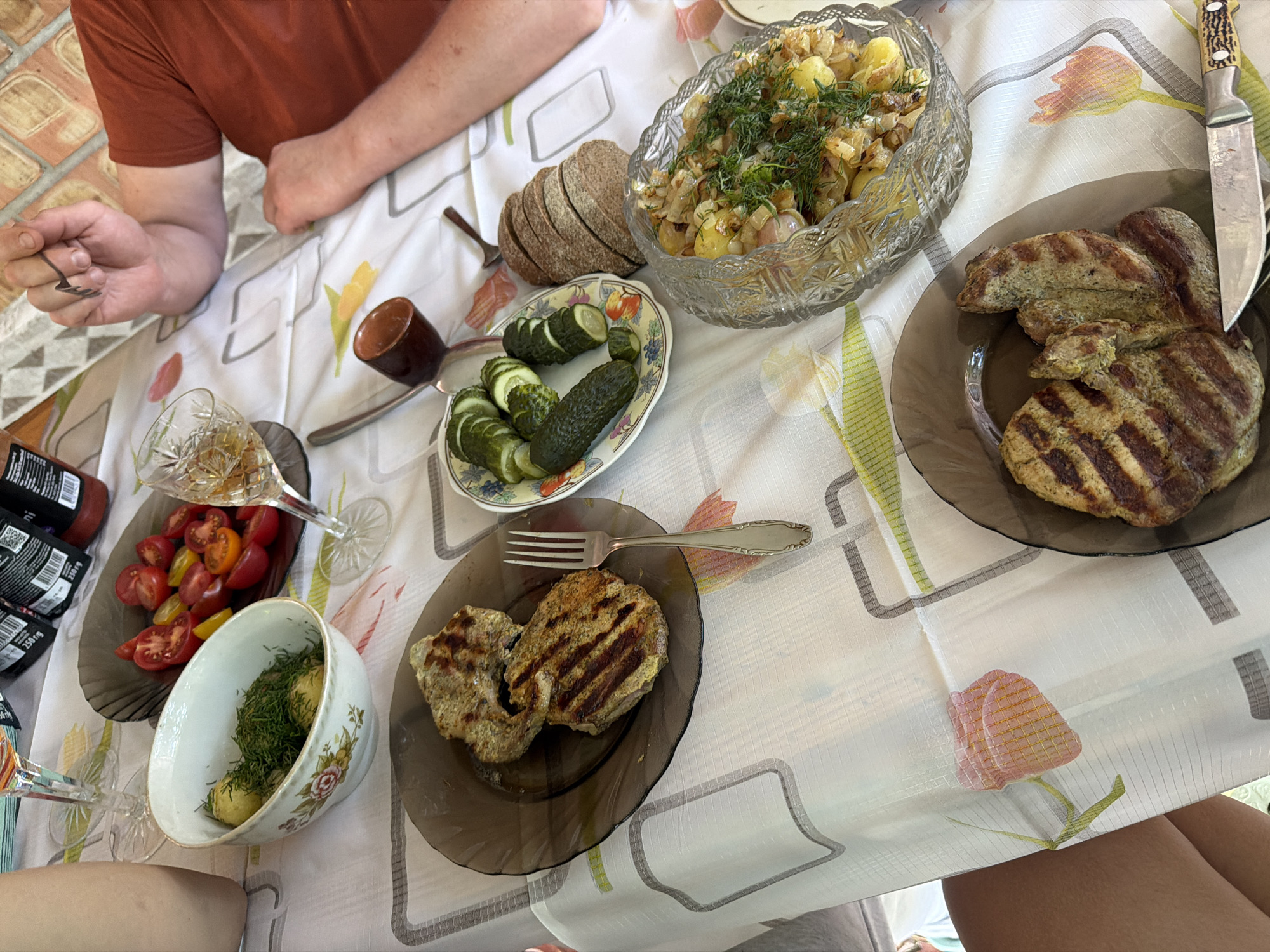 A table is laden with food and drinks, suggesting a meal is in progress or about to begin. The setting appears to be outdoors, likely a backyard or patio, given the dappled sunlight and the brick wall visible in the background, which is characteristic of some older buildings in Obukhivka, Ukraine.
On the table, there are several dishes. One plate contains two grilled meat patties, their surfaces marked with grill lines. Another plate holds sliced cucumbers, some whole and some cut into rounds. A bowl filled with boiled potatoes and fresh dill sits nearby. A crystal bowl contains a dish of potatoes and onions, garnished with dill. A small pile of sliced bread is also present.
Beverages include a glass with an amber liquid, possibly whiskey or brandy, and a small brown cup that could be for wine or another drink. Several bottles, some with labels suggesting they contain sauce or condiments, are also on the table.
Two individuals are partially visible. One person, wearing a rust-colored shirt, has their arm and hand resting on the table. Their hand is holding a small dark object, possibly a utensil or decorative item. The other person is only visible from the waist down, wearing a light-colored garment, with their legs crossed.
The table is covered with a white tablecloth featuring a floral pattern with stylized tulips and geometric lines. The overall impression is of a casual outdoor gathering for a meal.