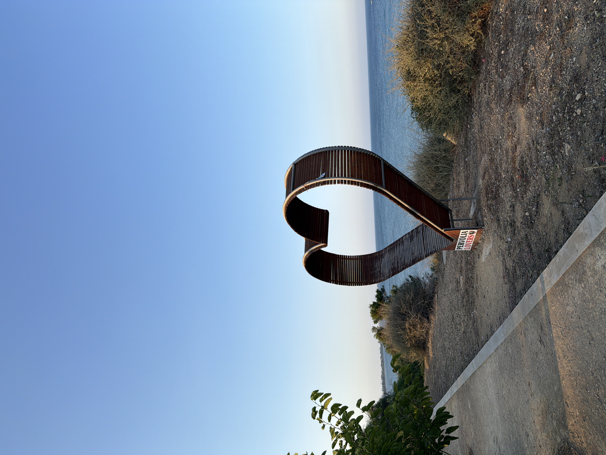 The image depicts a large, heart-shaped wooden sculpture positioned outdoors on a rocky, arid terrain overlooking a calm sea under a clear blue sky. The sculpture, constructed from curved wooden planks, faces the viewer with its opening directed towards the water. A small, red and white sign is affixed to the base of the sculpture, reading "PERVOLIA LOVERS." To the left of the sculpture, a paved walkway runs parallel to the foreground, bordered by dry earth and sparse vegetation. Lush green foliage is visible on the far left of the image. In the distance, the coastline stretches out, with the sea meeting the horizon. The overall scene suggests a seaside location, likely a public space or park, in Pervolia, a village within the municipality of Meneou, Cyprus. There are no people visible in the frame. The lighting indicates it is either early morning or late afternoon.