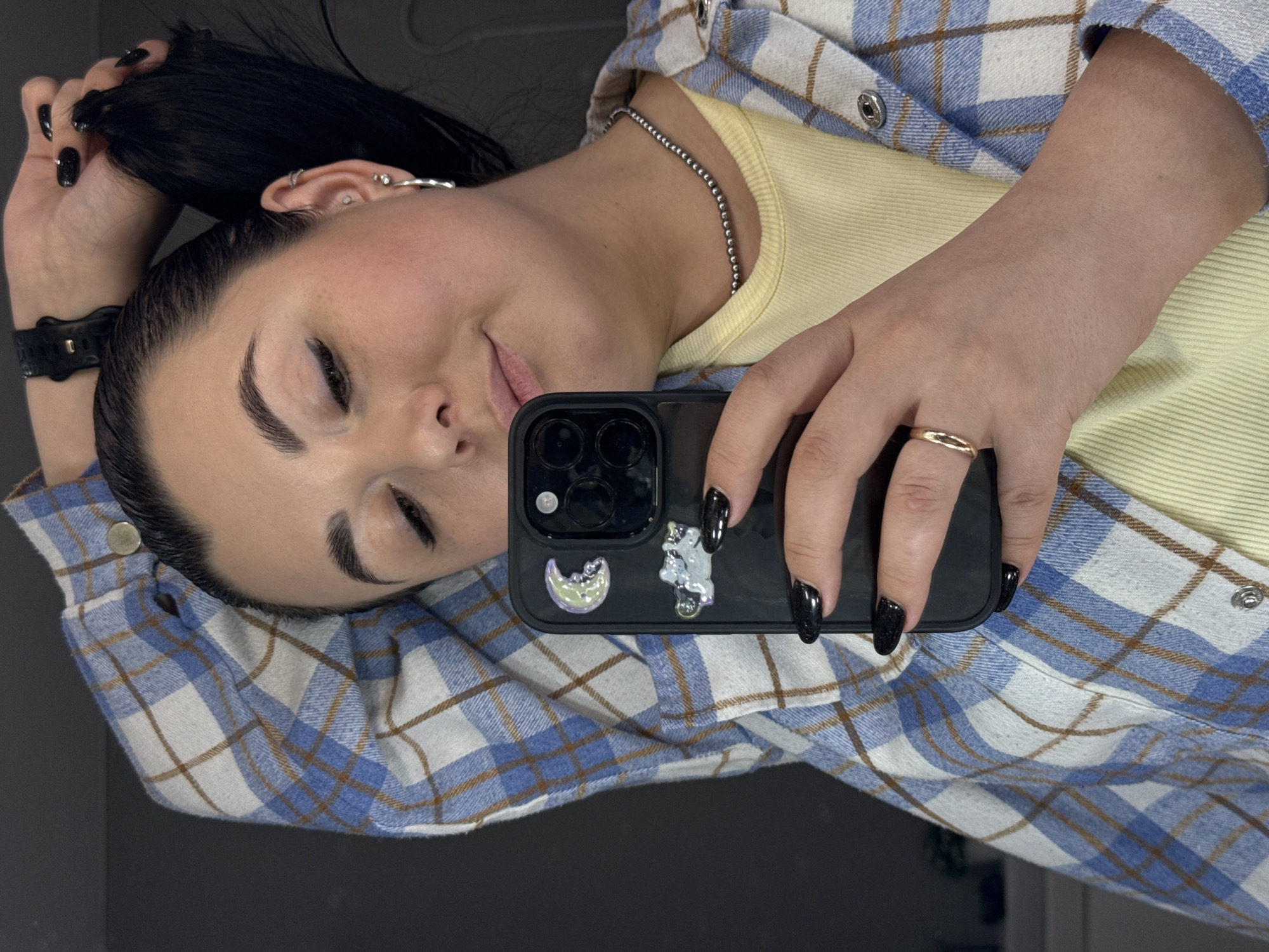 A young woman is captured in a close-up, laid-back selfie, seemingly taken indoors. She has dark hair, pulled back, with a few strands held by her left hand. Her eyebrows are well-defined, and her eyes are partially closed, conveying a relaxed expression with a subtle smile.
She is wearing a light yellow, ribbed-knit top layered under a blue, white, and brown plaid flannel shirt. Her accessories include a thin silver beaded necklace, multiple silver earrings/piercings on her left ear, and a black digital watch on her left wrist. On her right hand, she wears a simple gold band ring on her ring finger, and her fingernails are painted with black polish.
She holds a black smartphone, visible in the foreground, with its reflective screen showing a glimpse of her face. The phone case features two decorative, iridescent stickers: one depicting a crescent moon-like shape and another an abstract figure. The background is a plain, dark surface, suggesting an uncluttered indoor setting. The image is rotated, showing her lying horizontally. There is no visible text in the image.