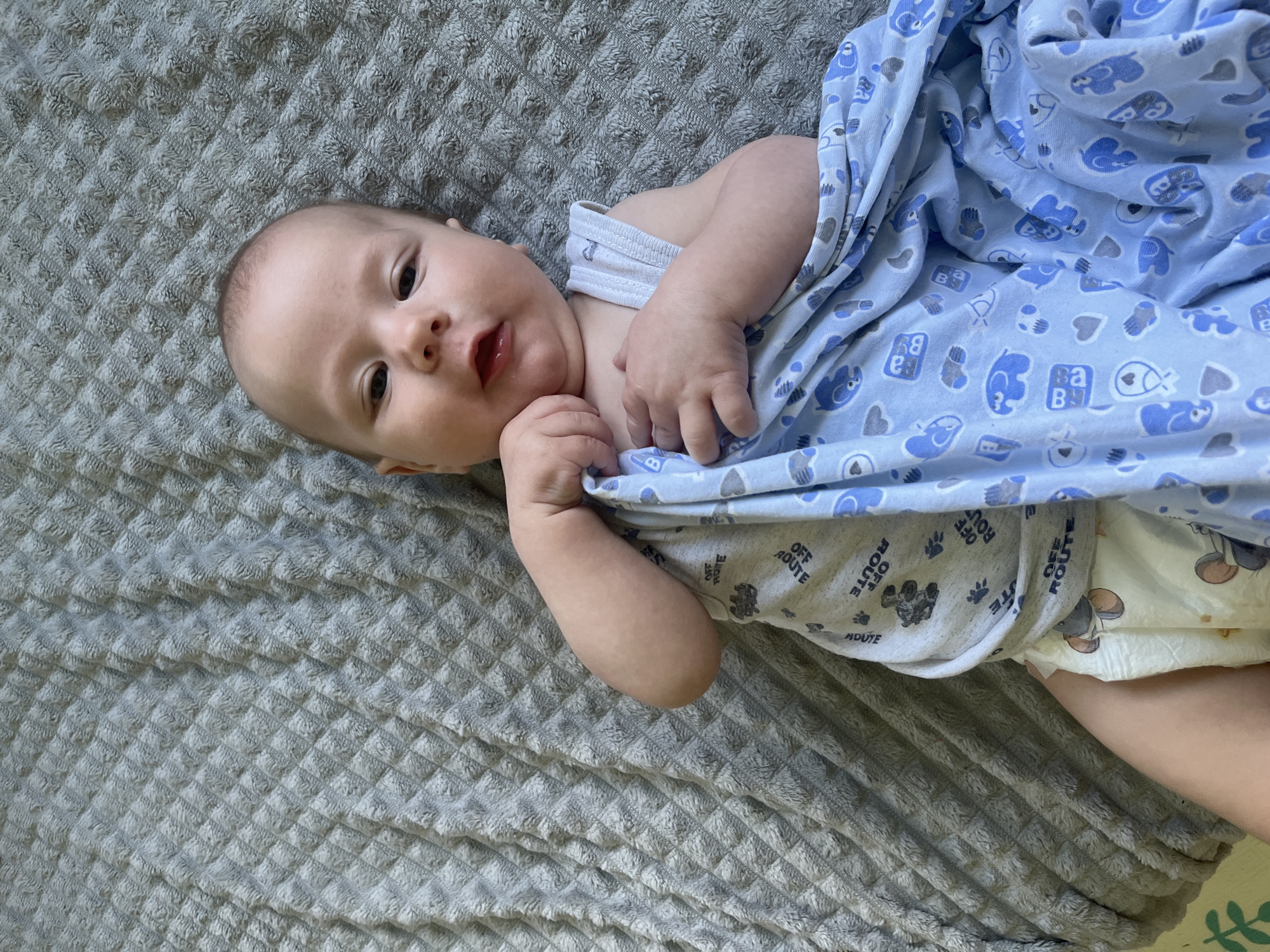 A baby is lying on a grey textured blanket. The baby is wearing a grey onesie with a black bear print and the words "OFF ROUTE" repeated, and a diaper with a cartoon print. The baby is partially covered by a blue blanket with a pattern of elephants, hearts, and the letters "B A B". The baby's head is turned towards the camera, with its mouth slightly open. The baby's hands are near its chest. The background is entirely the grey blanket. This image was taken in Andreyevo, Russia.