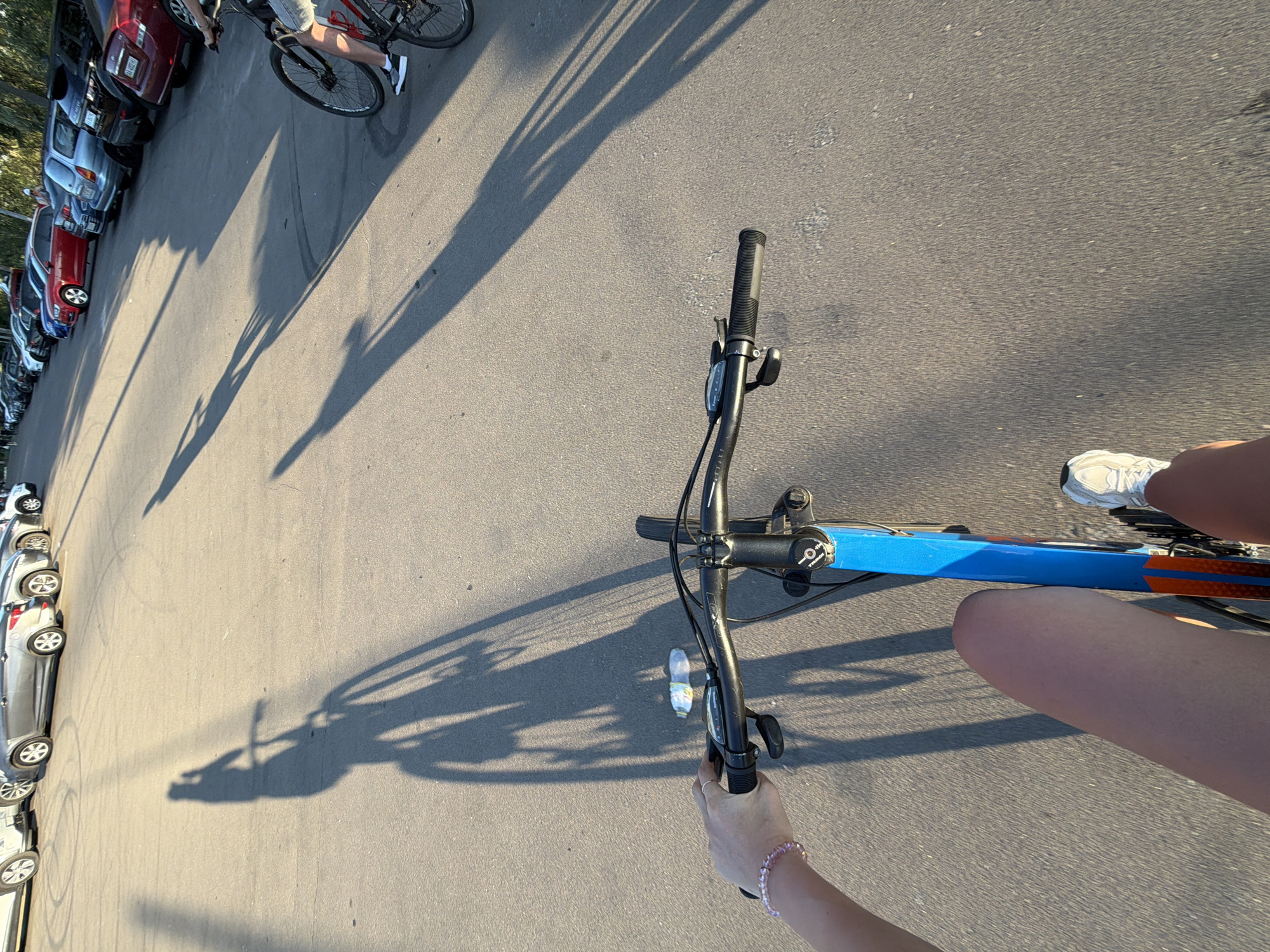The image depicts a first-person perspective of someone riding a blue and orange bicycle on an asphalt surface, likely a parking lot or street, in Kyiv, Ukraine. The sun is low, casting long shadows from the bicycle and rider across the ground. In the background, a row of parked cars is visible, including a red sedan and several SUVs. The rider's hand, adorned with a pink beaded bracelet, is on the handlebars, which feature gear shifters and brake levers. A partially visible water bottle lies on the ground near the bicycle. The rider is wearing athletic shoes and shorts. Another cyclist and their bicycle are visible further down the row of cars on the right side of the frame.