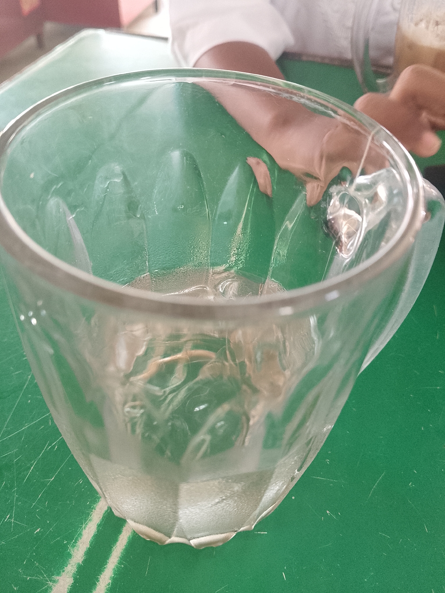 The image shows a clear, empty glass mug with a faceted design sitting on a green surface. The mug has a handle on the right side. In the background, there is a person wearing a white shirt. Another mug, containing a light brown beverage, is partially visible in the upper right corner. The green surface appears to be a table, marked with white lines. Based on the prompt of Lhokseumawe, Indonesia, the scene likely takes place in a cafe or restaurant there.
