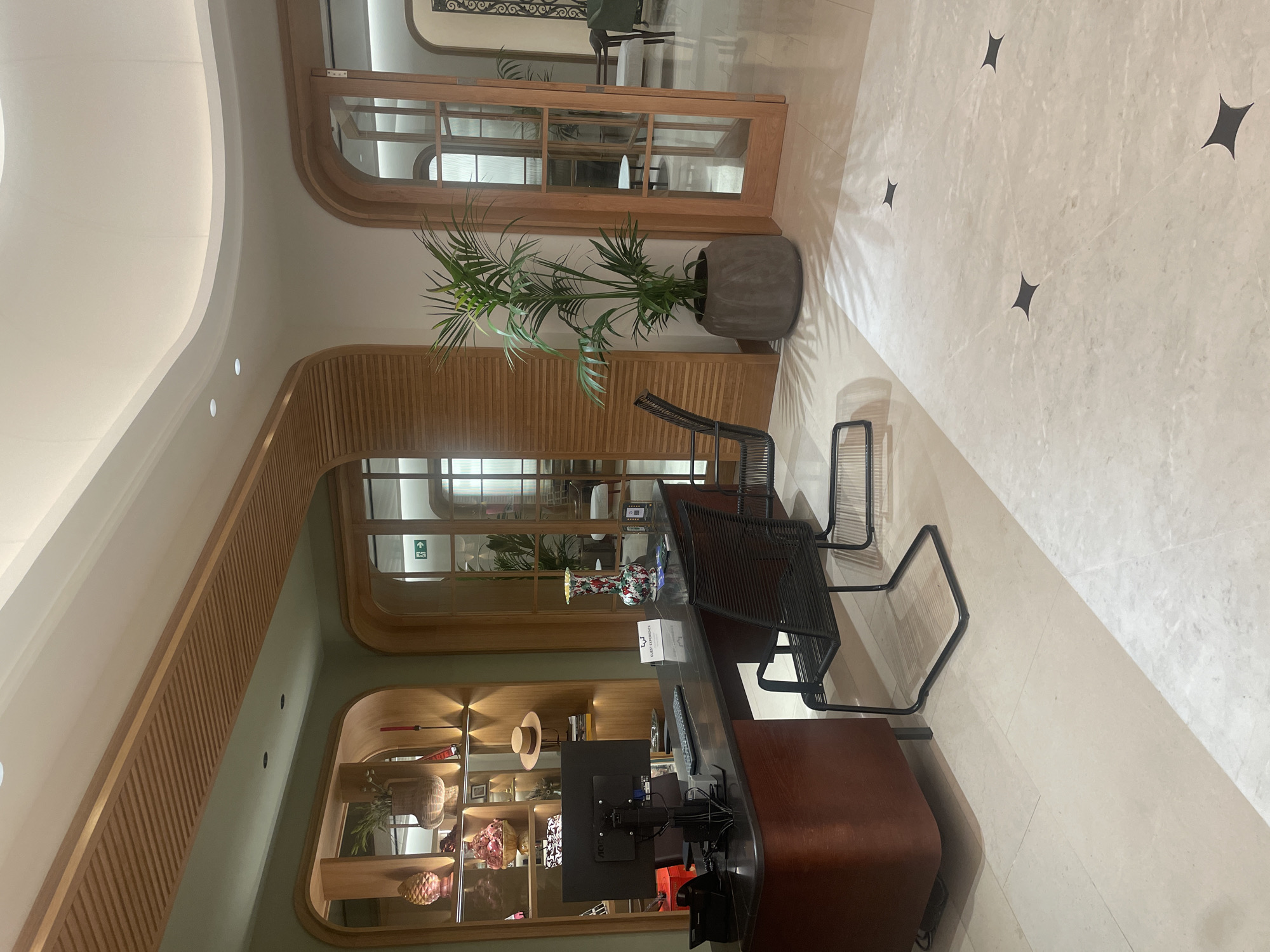 The image shows the reception area of a hotel in Seville, Spain. The reception desk is made of dark wood and marble, with a computer, keyboard, mouse, telephone, and a vase of flowers on it. There are two chairs in front of the desk. To the left of the desk, there is a wall with shelves displaying various decorative items, including a pineapple, a woven basket, and a hat. To the right of the desk, there is a large potted plant. In the background, there are two sets of double doors made of wood and glass, leading to other areas of the hotel. The floor is tiled with large light-colored tiles. The overall ambiance is modern and elegant.