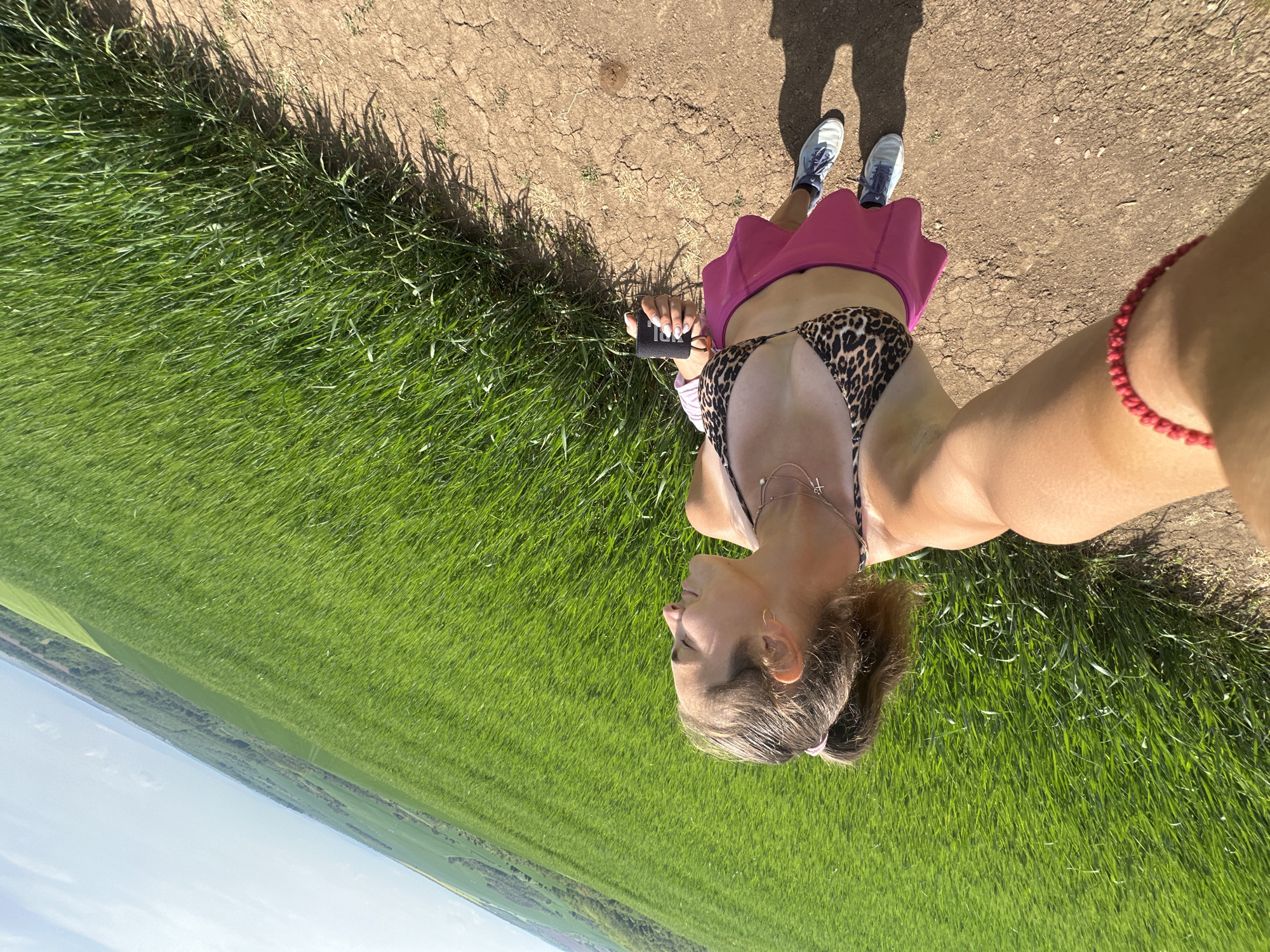A person wearing a leopard-print bikini top and pink skirt stands on a dirt path beside a field of green grass. The person is holding a small black object in their left hand and wearing a red bracelet on their right wrist. They are also wearing light-colored athletic shoes. The background shows a distant horizon line and sky. The image appears to be taken from a high angle, possibly a selfie. No information indicates the location is Kashin, Bulgaria.