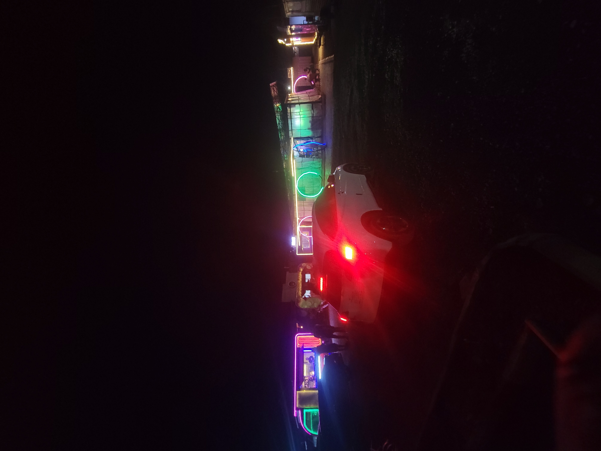 The image shows a brightly lit roadside establishment in Jos, Nigeria at night. The establishment consists of several connected stalls or structures adorned with neon lights of various colors, including green, blue, pink, and yellow. A white car is parked beside one of the stalls, with its red taillights illuminated. People are present near the stalls, but their features are obscured due to the darkness. The environment is dark, with the only visible details being the bright lights of the establishment and the car. Vegetation is visible in the background.