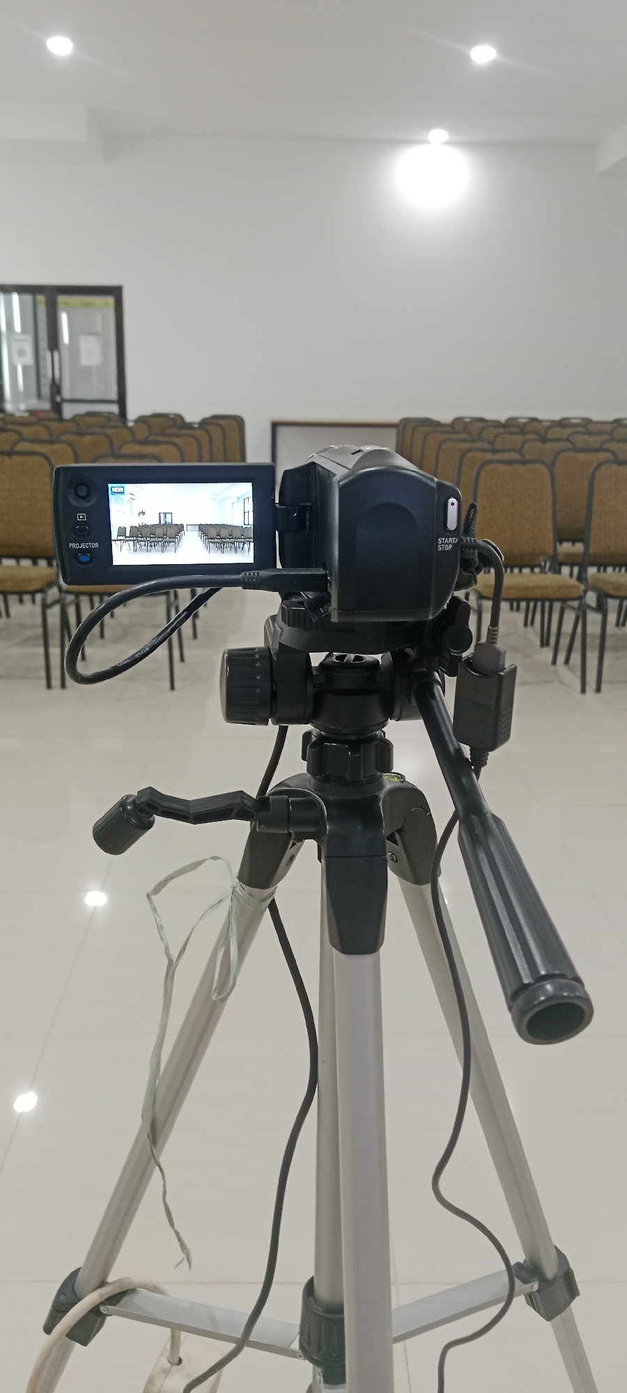 The image shows a video camera mounted on a tripod in what appears to be an indoor venue. The camera's screen displays a view of rows of empty chairs, suggesting a conference room, hall, or auditorium. The background consists of more rows of chairs, and a white wall with ceiling lights. The floor is tiled. The scene is well-lit, likely by artificial lighting. The camera is positioned to record an empty seating area, possibly in anticipation of an event. The overall impression is one of preparation or a pause before an activity. The city of Tuban, Indonesia, is not explicitly indicated in the image, but the context suggests a public or semi-public indoor space typical of meeting or event venues in such a location.