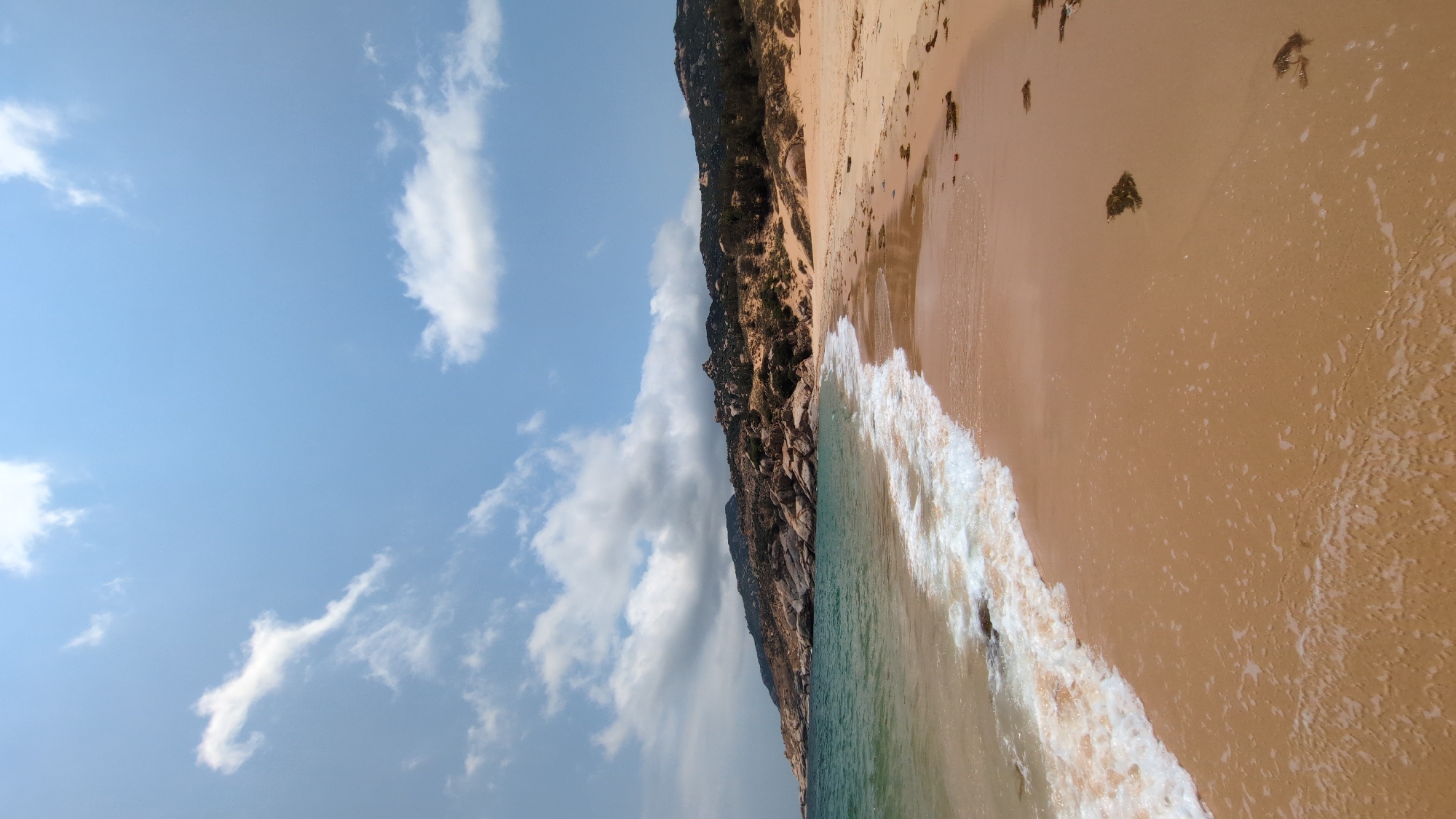 A shoreline, likely in Phước Dinh, Vietnam, is depicted in this high-angle, outdoor shot. The image is rotated counter-clockwise.
The left side of the frame is dominated by a bright blue sky with scattered, puffy white clouds. On the right is a sandy beach.
A line of dark, rocky cliffs separates the sky from the shoreline. The cliff face is steep, vegetated, and appears rugged.
The sandy beach stretches from the cliff base towards the right edge of the image. The ocean's waves reach the shore. The water is a mix of blue and green hues. The wave edges are lined with white foam. Small clumps of seaweed are visible on the sand.