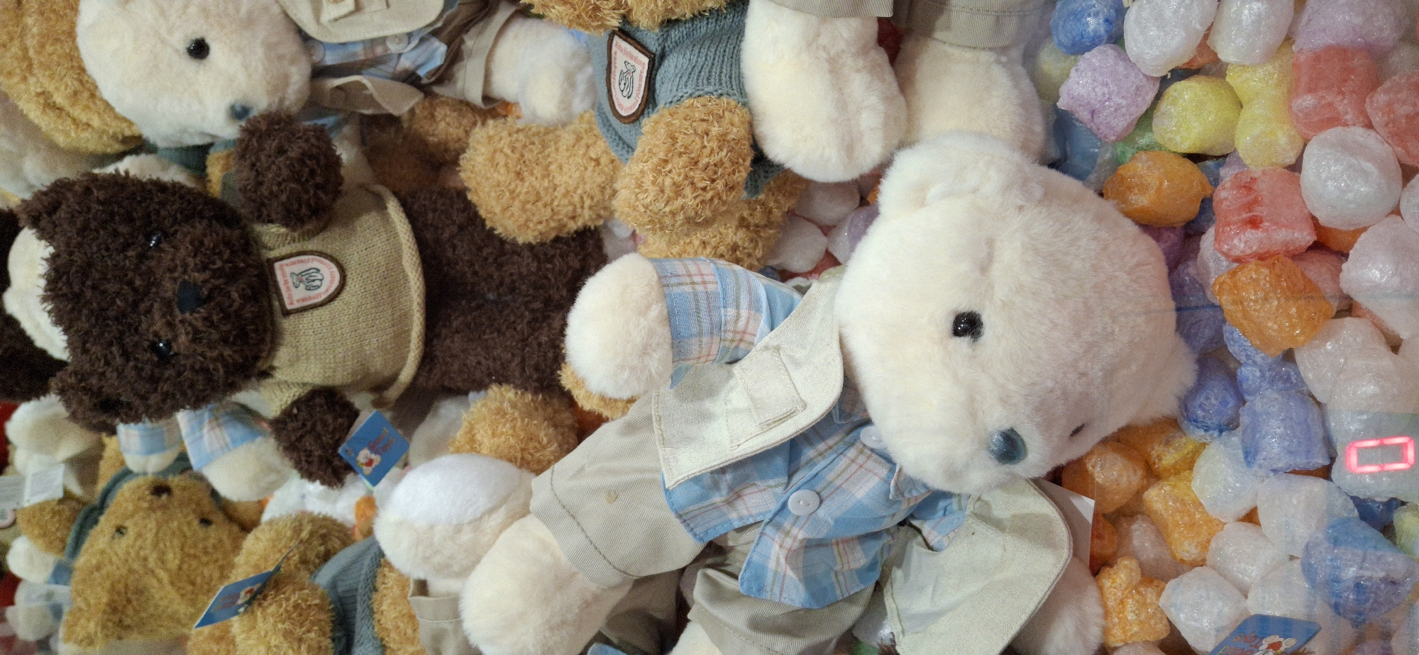 A close-up, overhead shot depicts a pile of stuffed teddy bears and translucent, brightly colored, irregularly shaped objects. The teddy bears vary in color and size, with some wearing small sweaters and jackets. One teddy bear in the foreground, facing left, has a white head and dark eyes. In the background, a dark brown teddy bear wears a tan sweater with a crest. The colorful objects appear to be small, translucent pieces of plastic or candy. The scene is illuminated by artificial light. This image was captured in the city of Telmana, Russia.