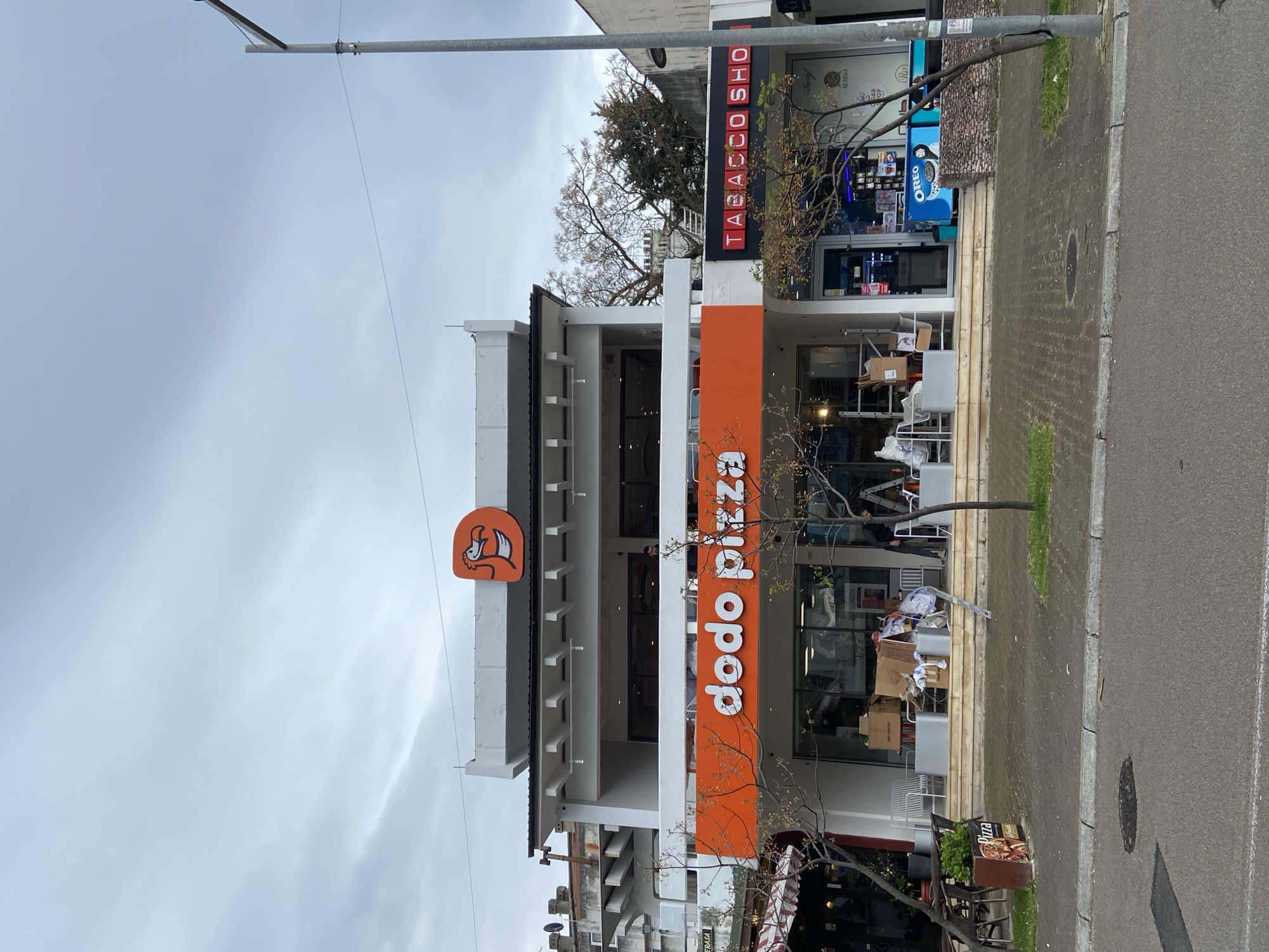 A ground-level, daytime photograph depicts a Dodo Pizza restaurant located in Bar, Montenegro. The restaurant's signage, prominently displayed in orange with white lettering, is visible. The building has a multi-tiered white facade. A smaller "Tobacco Shop" is visible adjacent to the pizza establishment. An Oreo branded vending machine is in front of the Tobacco Shop. The sky is overcast. A streetlight is on the left side of the photo. The ground is paved, possibly a sidewalk and street. There is some outdoor seating on the terrace. There is what looks to be a pile of trash next to some outdoor seating. The photo shows a small portion of a tree on either side of the image.