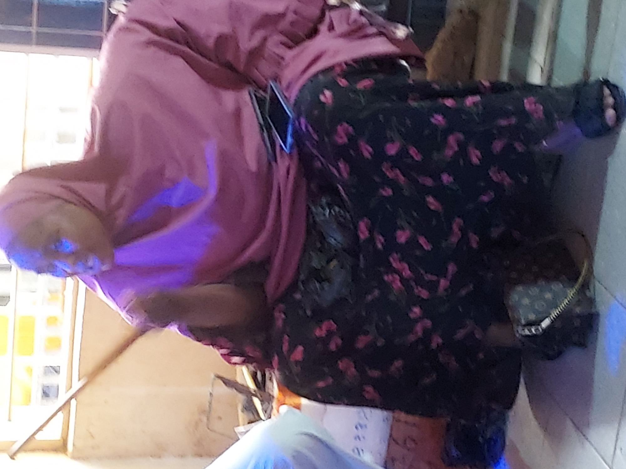 An indoor image, rotated clockwise, features a person dressed in a vibrant pink or magenta-colored long garment, possibly a loose top with a head covering, and dark pants adorned with a scattered pink floral pattern. A distinct blue-purple light illuminates the person's head and upper body, creating a glow.
A smartphone is visibly tucked into the side of the person's garment, and a small, light-colored handbag or pouch with a metallic zipper can be seen near their right hip. The background consists of a light-colored wall and what appears to be a window or glass partition with a grid pattern, allowing some external light into the space. The floor is a light beige or brown.
The person appears to be standing or posing casually. There is no clear activity taking place. The prominent artificial blue-purple light suggests the photograph may have been taken in the evening or a dimly lit indoor environment. Faint and illegible text is visible on some papers or a book in the lower section of the image. While the image itself offers no specific geographical markers, it is provided with the location context of Jos, Nigeria.
