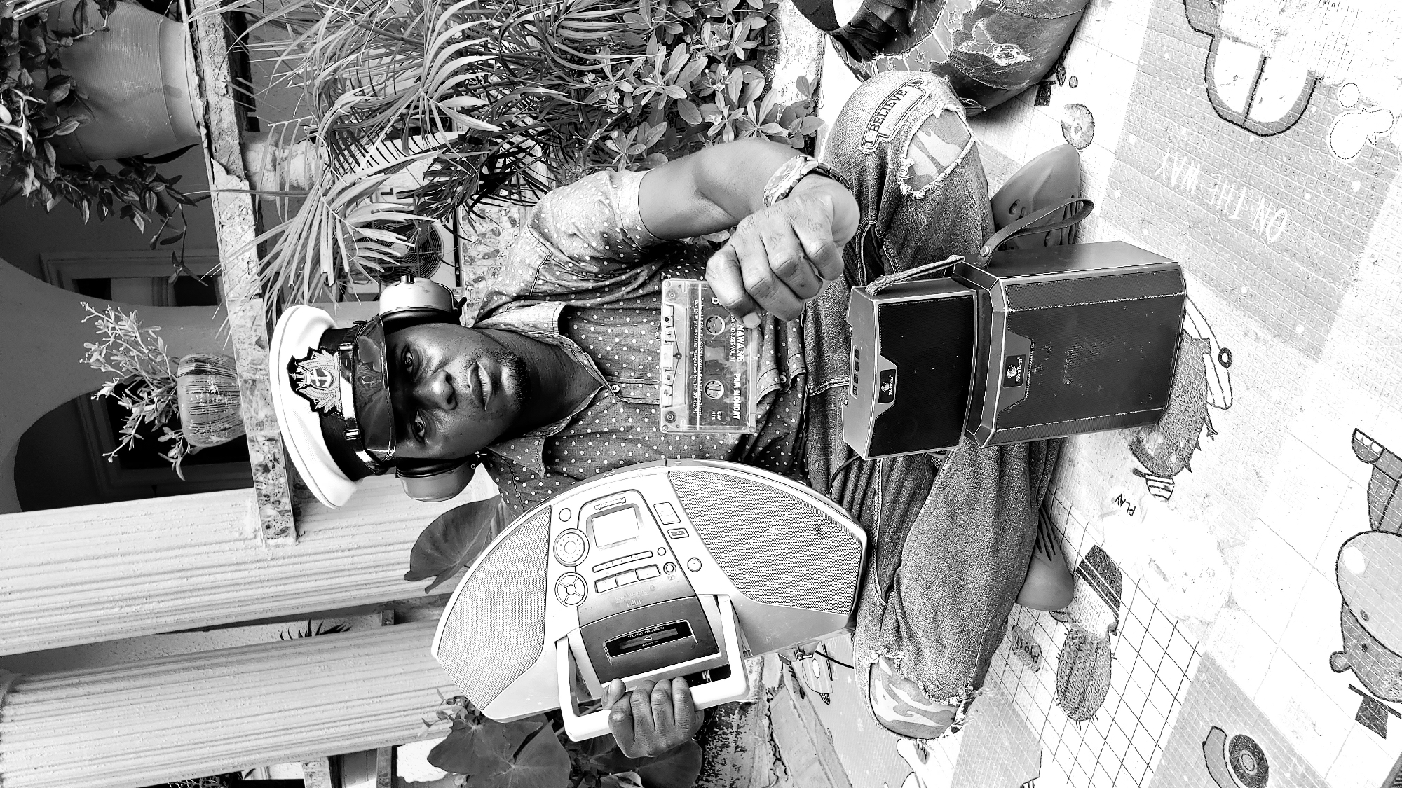 A black and white, medium-shot photograph captures a Black male seated outdoors, outdoors in the city of Narayi, Nigeria, on a patterned mat. The man, wearing a naval-style hat and headphones, holds a boombox in his left hand and a cassette tape in his right. He is dressed in a polka-dot collared shirt and ripped jeans, with two black speakers stacked in front of him. In the background, large pillars and lush green foliage suggest a tropical or sub-tropical environment, common in parts of Nigeria. The scene is likely a domestic or public outdoor space, with the overall ambiance hinting at a nostalgic or retro theme, possibly related to music and older technology.
